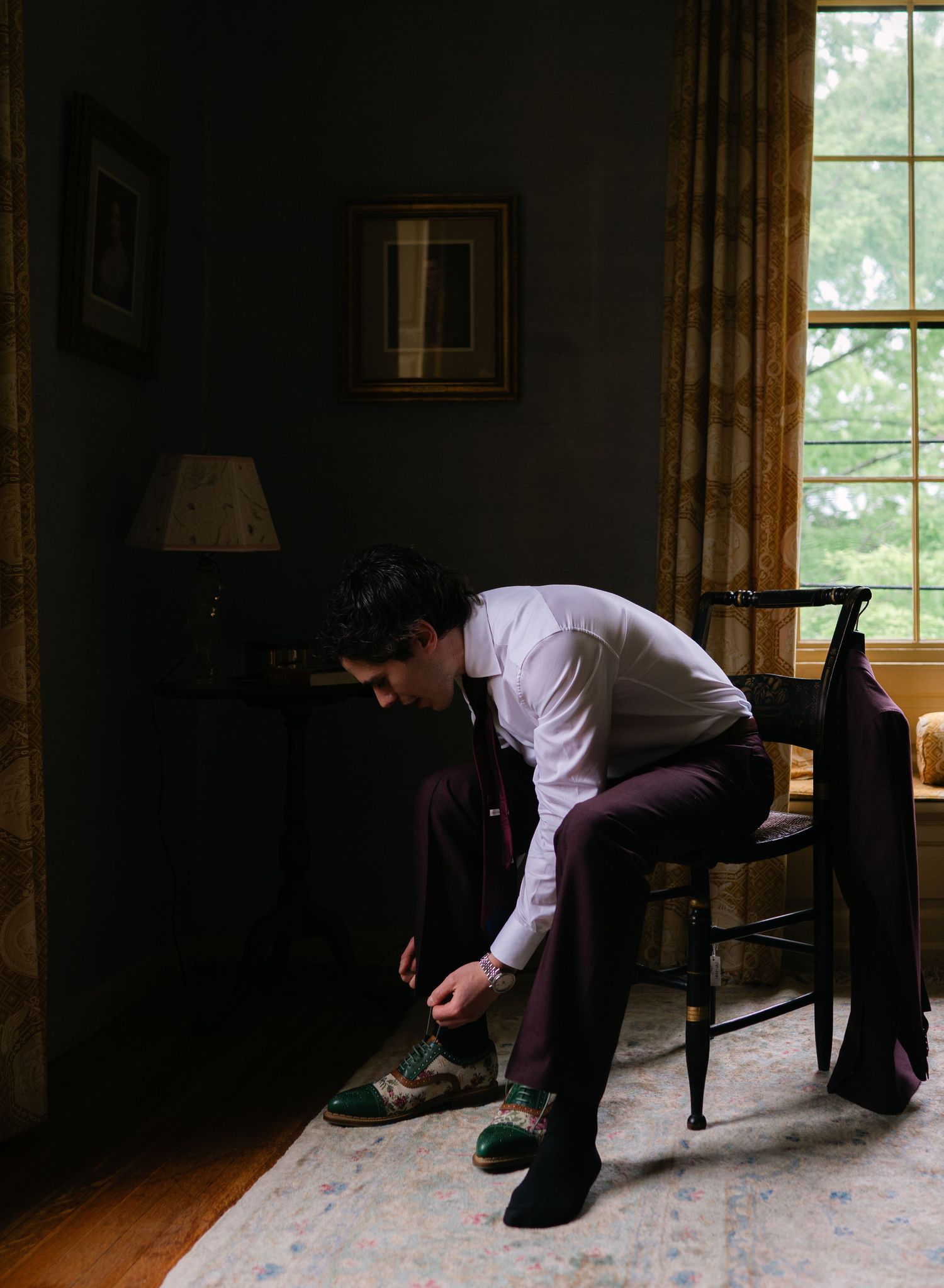 A groom in burgundy suit ties his shoes on a wooden chair in a dimly lit room with a window.