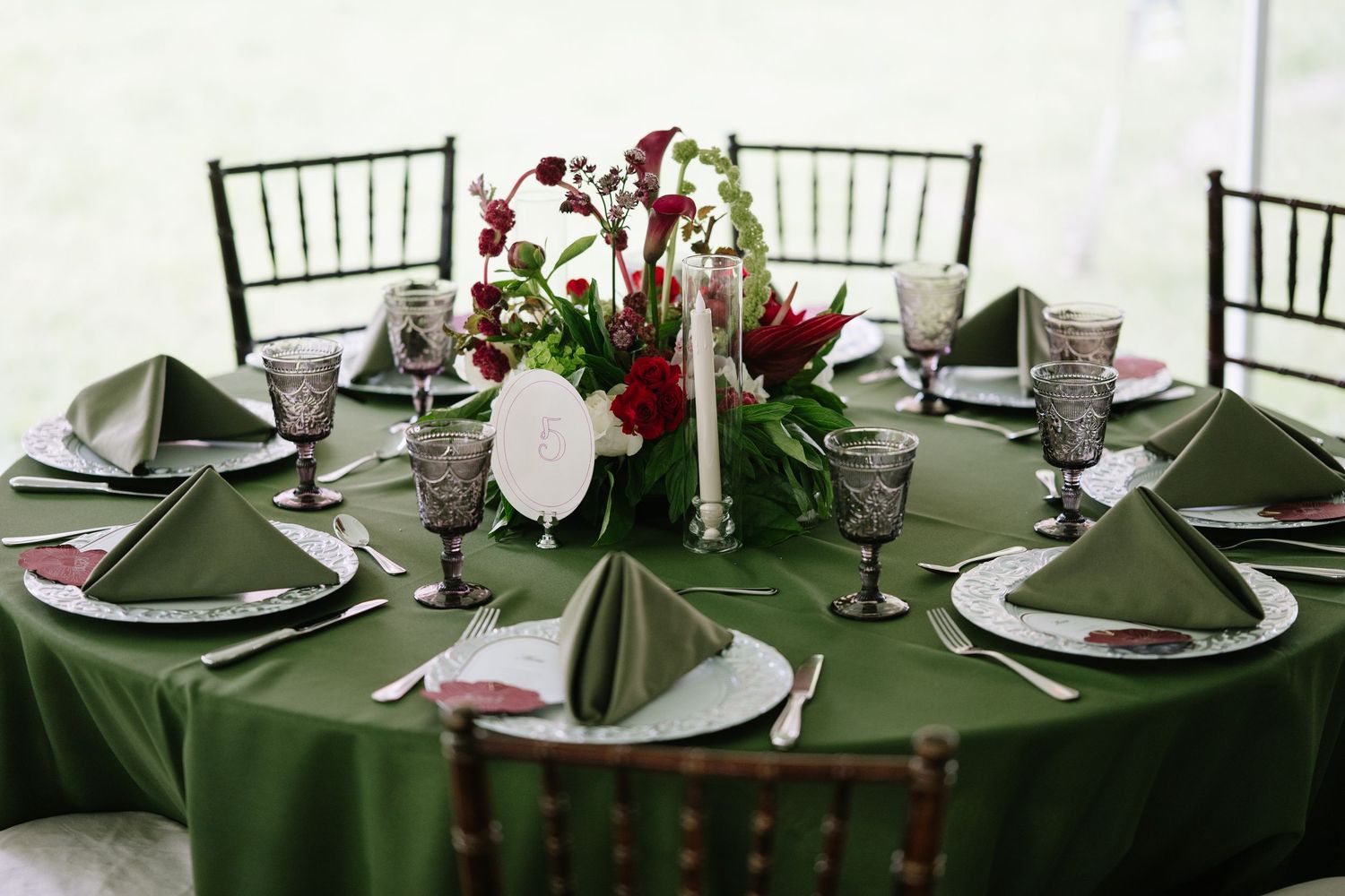 Elegant green tablecloth round table with dark wood chairs, floral centerpiece, gray goblets, and folded green napkins.