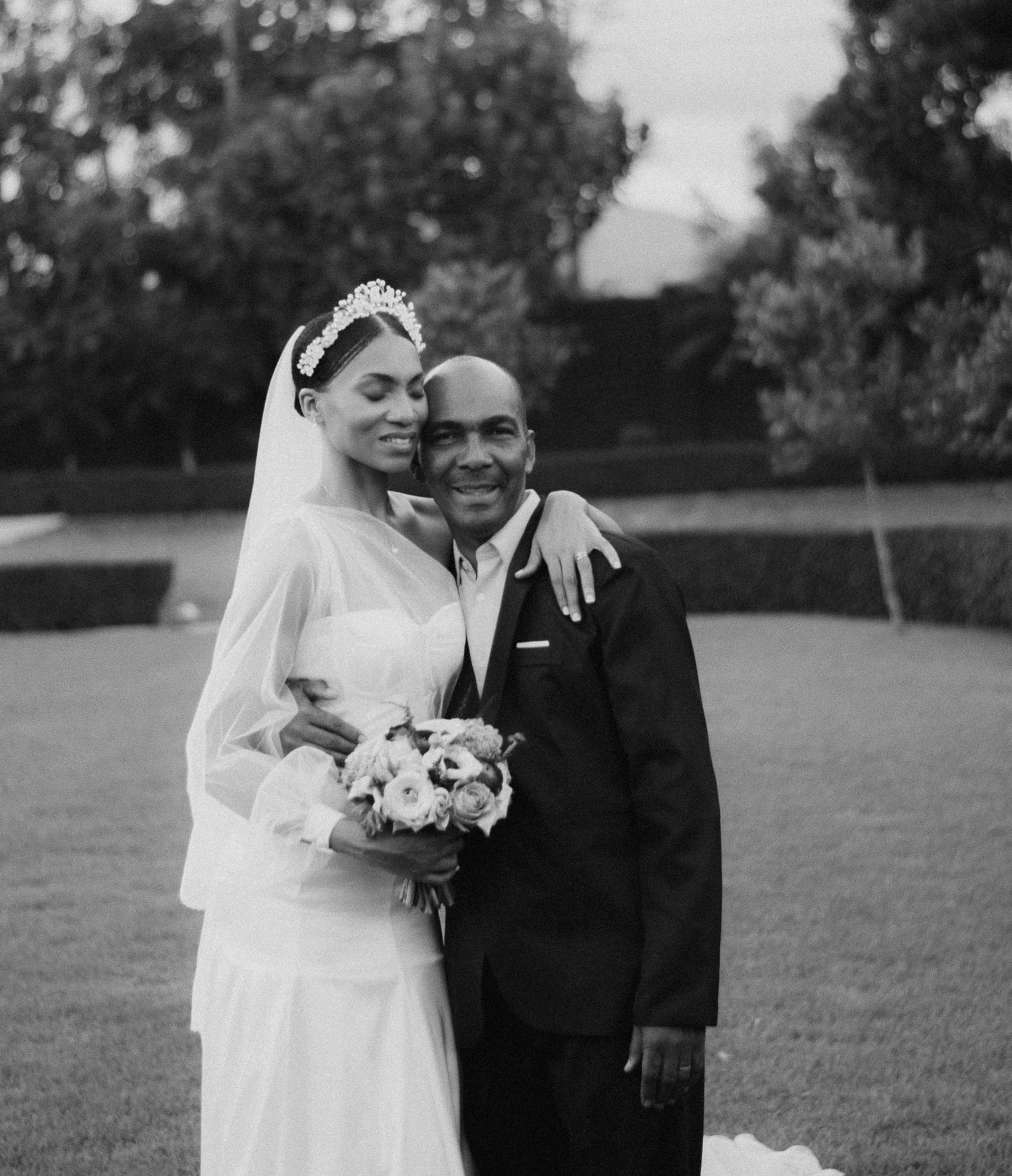 A romantic black and white outdoor wedding portrait of a couple embracing on a lawn with trees in background.