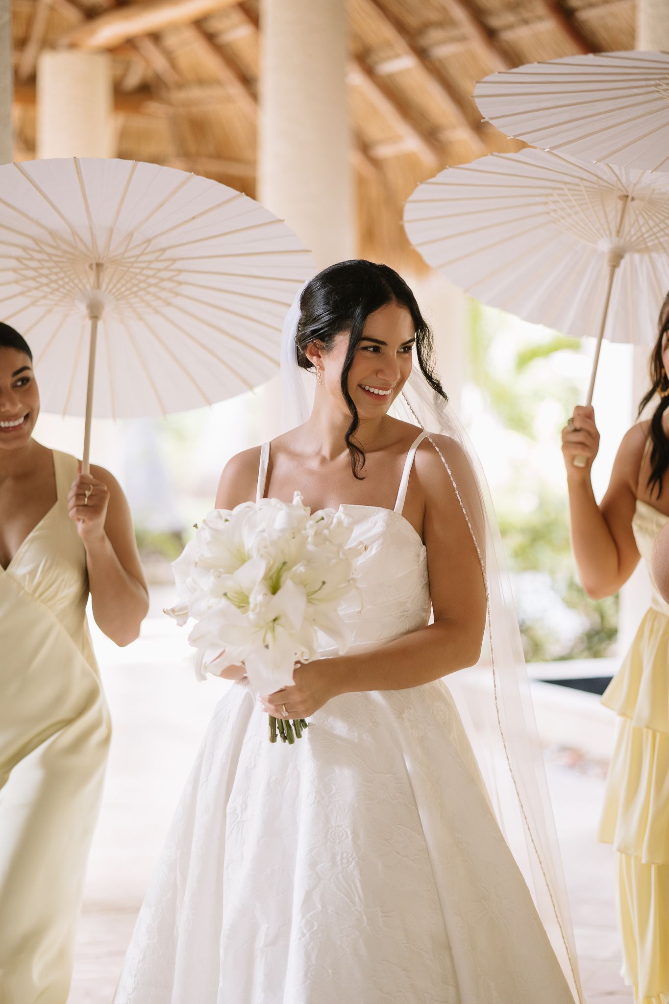 Bridesmaids in light yellow dresses holding white parasols pose together outdoors.