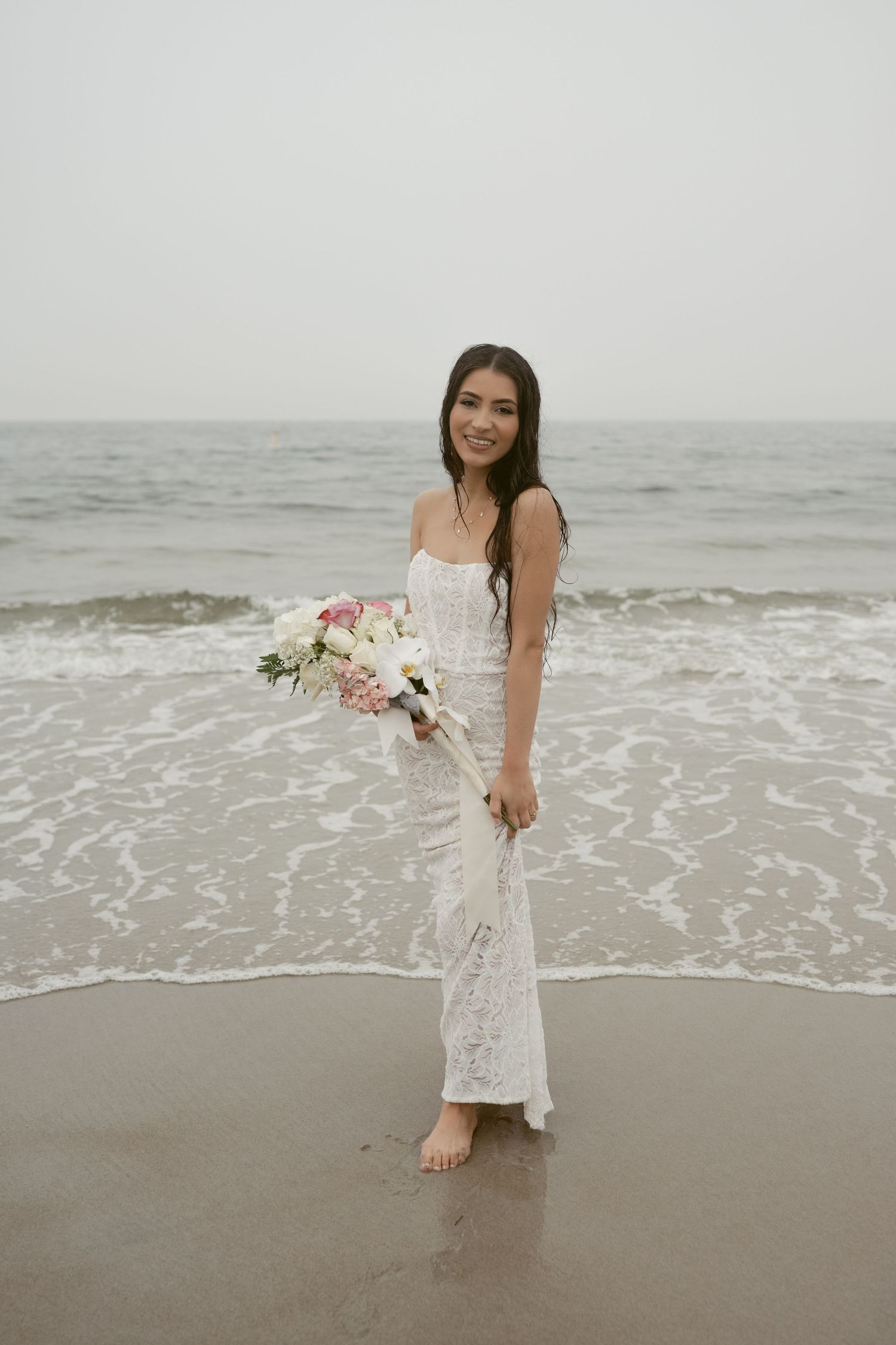 A bride in a white lace dress plays in the ocean waves during a beach photoshoot.