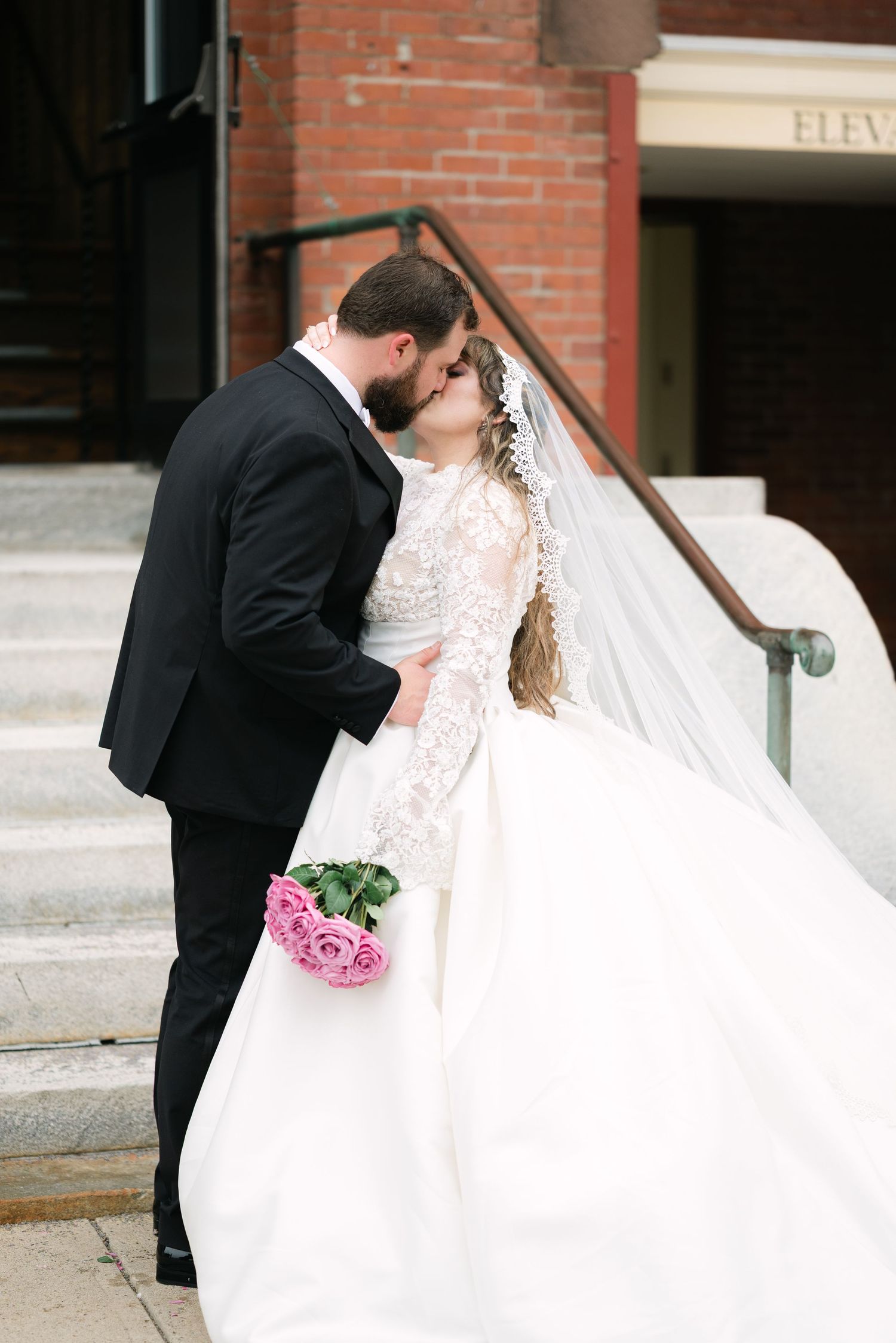Bride in lace dress with flowing veil shares romantic moment outside brick building.