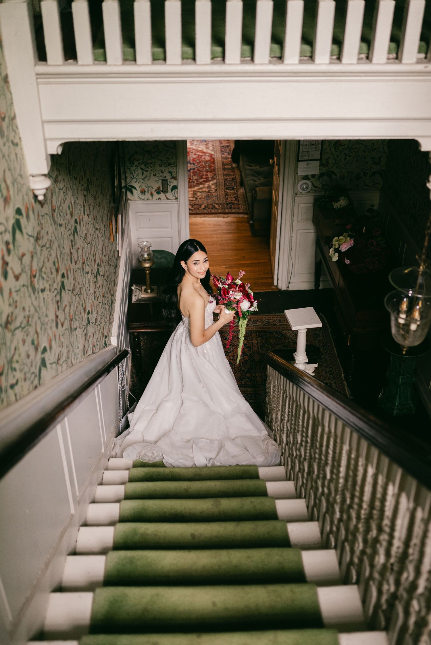 Bride in white gown holding red bouquet poses on green-carpeted staircase in elegant vintage venue.