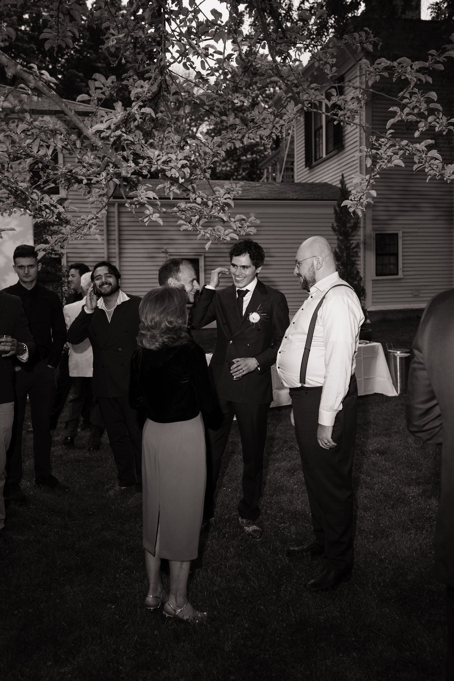 Black and white photo of wedding guests conversing outdoors, groom in suit with boutonniere, backyard reception setting.