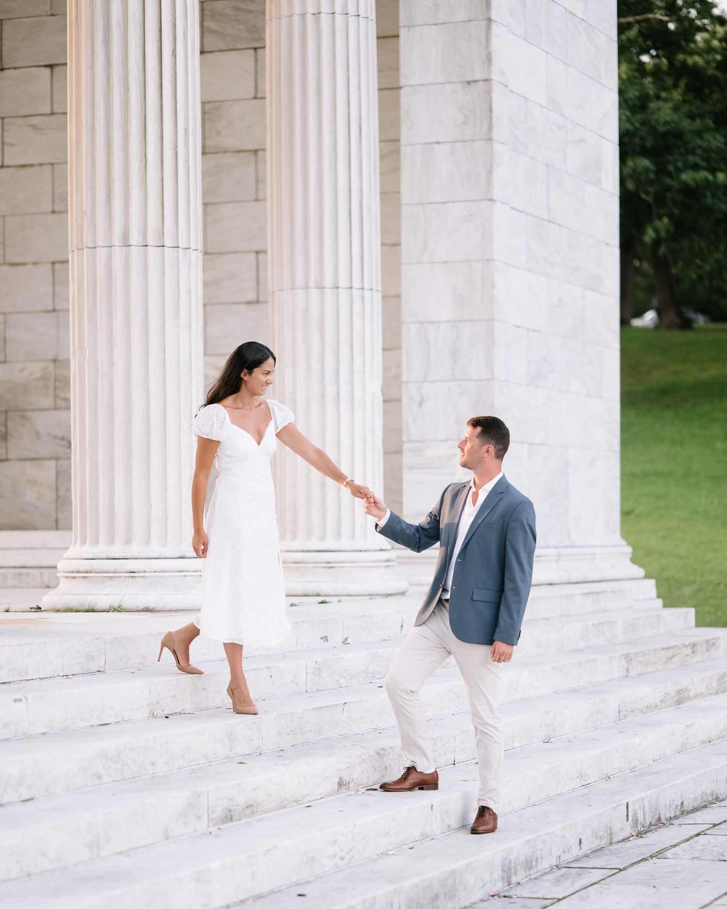 Couple poses playfully on white stone steps surrounded by classical columns.