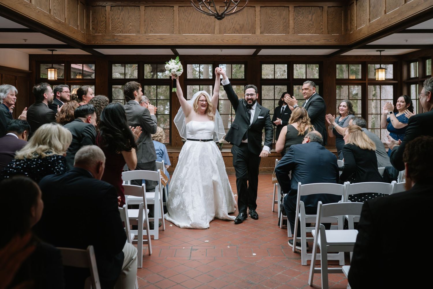 Bride and groom raising hands in joy at wedding ceremony aisle, guests applauding in elegant wood-paneled venue.
