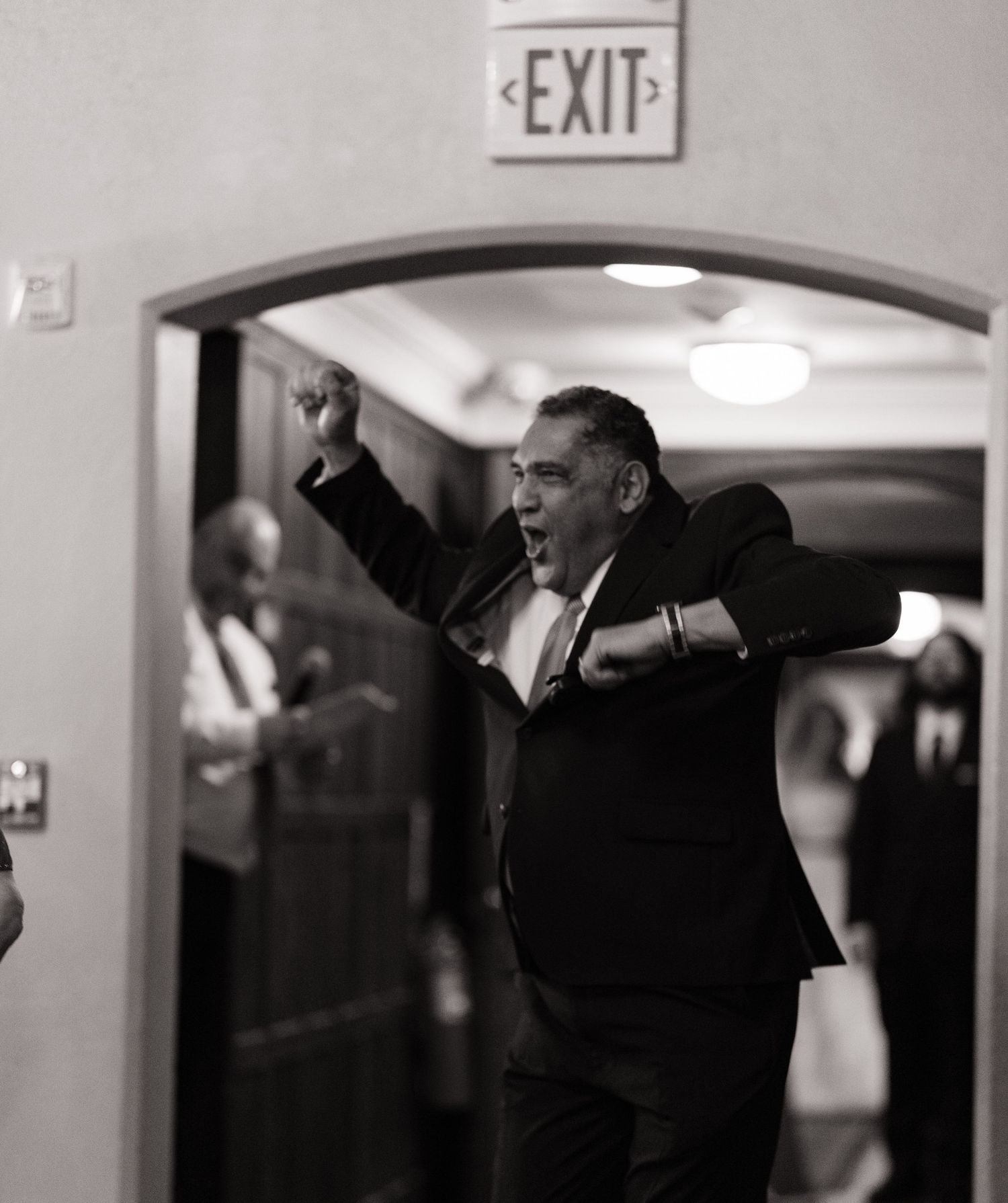 Black and white photo of an excited man in a suit raising his fist under an EXIT sign at a wedding reception.