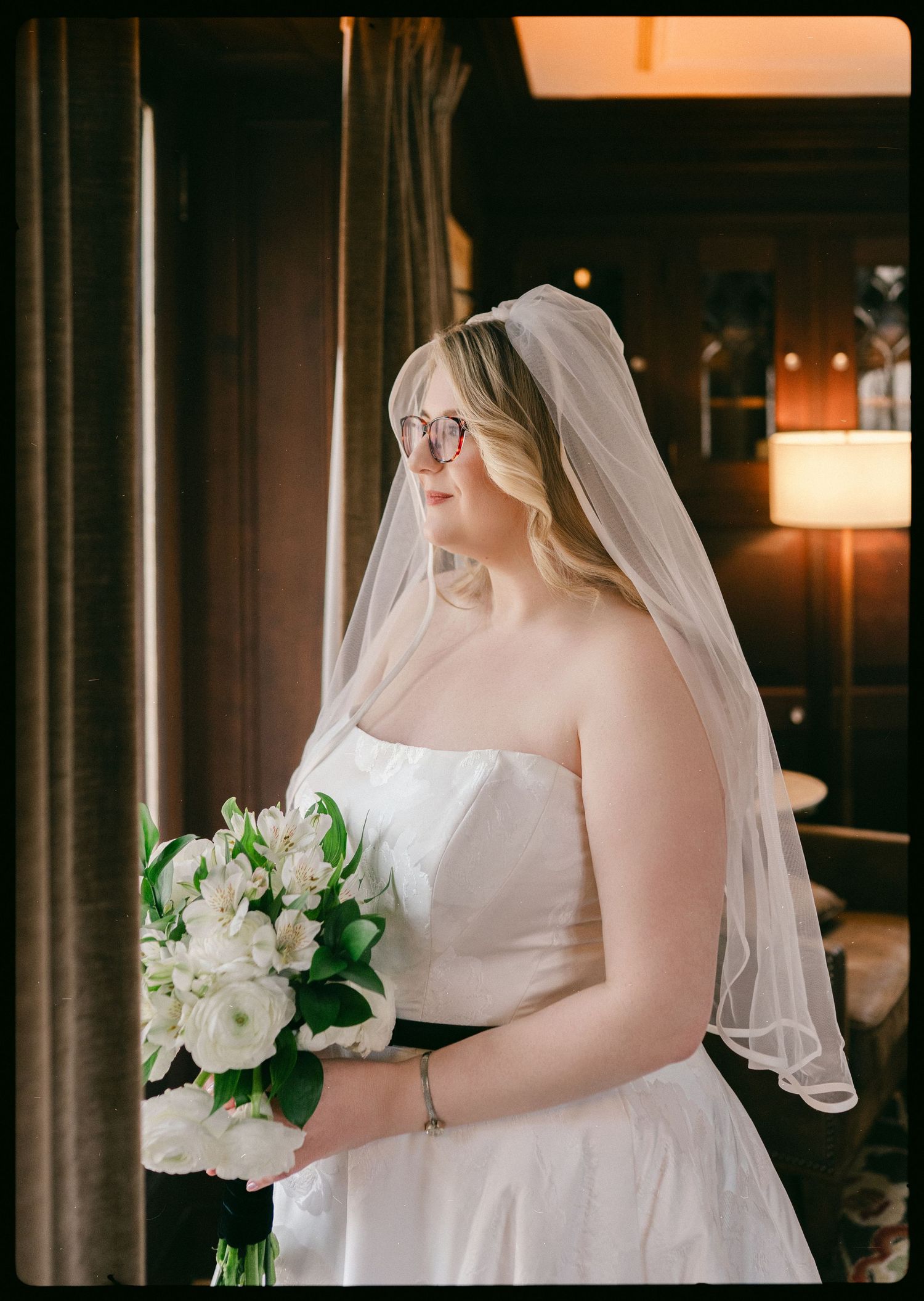 Bride in strapless white gown and veil holding white floral bouquet, wearing glasses, standing by window in elegant venue.