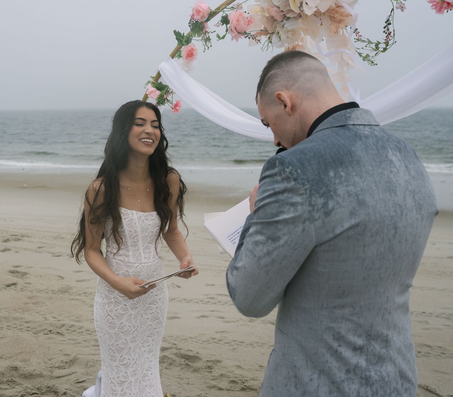 A romantic beach wedding photoshoot with the couple laughing and embracing as a white veil flows in the ocean breeze.