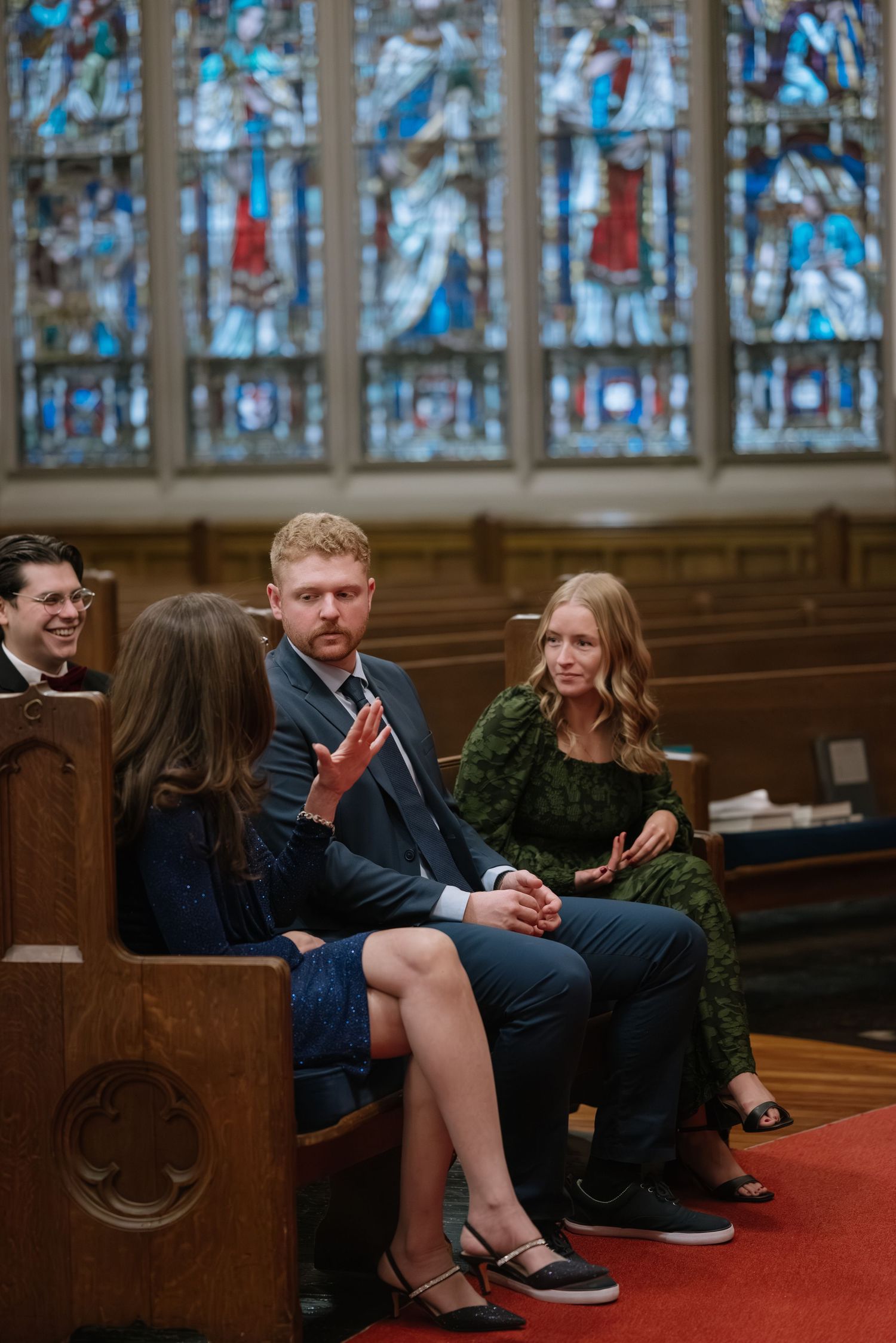 People having an animated conversation in church pews beneath stained glass windows.