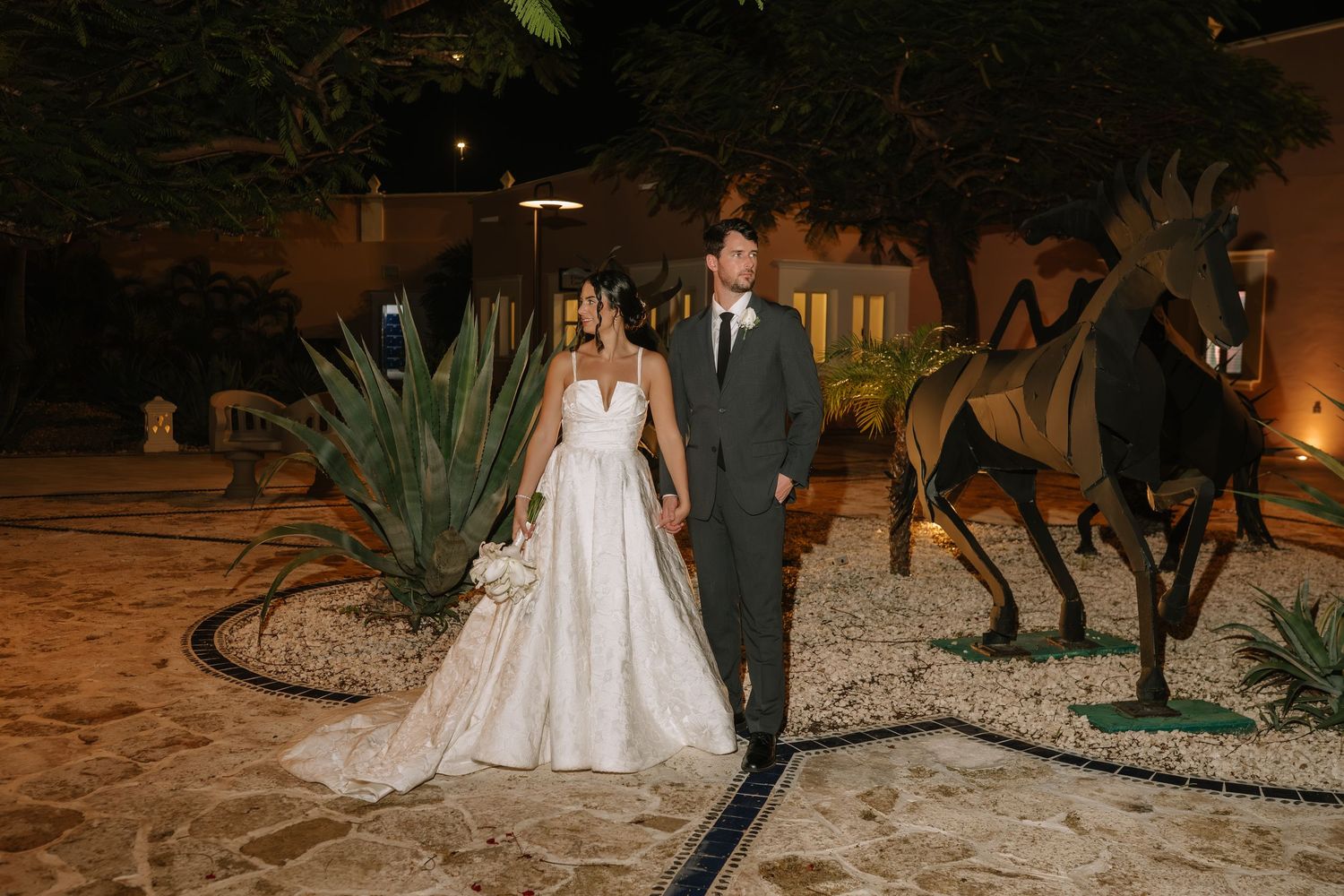 A couple in wedding attire stands together at night in a garden setting with decorative metal sculptures.