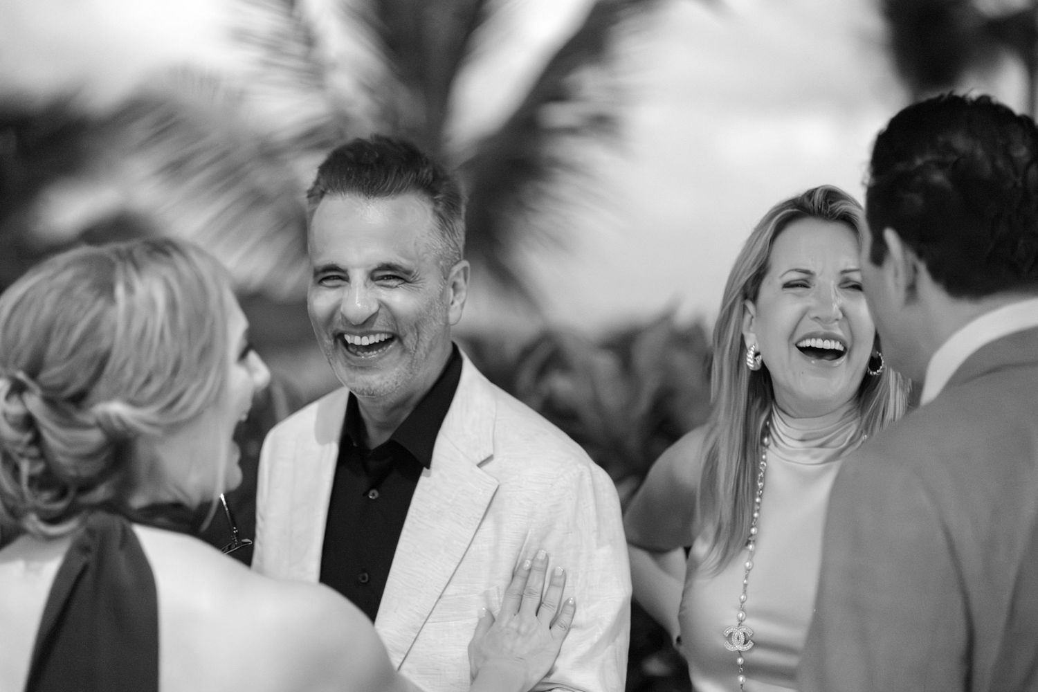 Black and white photo of people laughing and socializing at an outdoor event with palm trees in background.