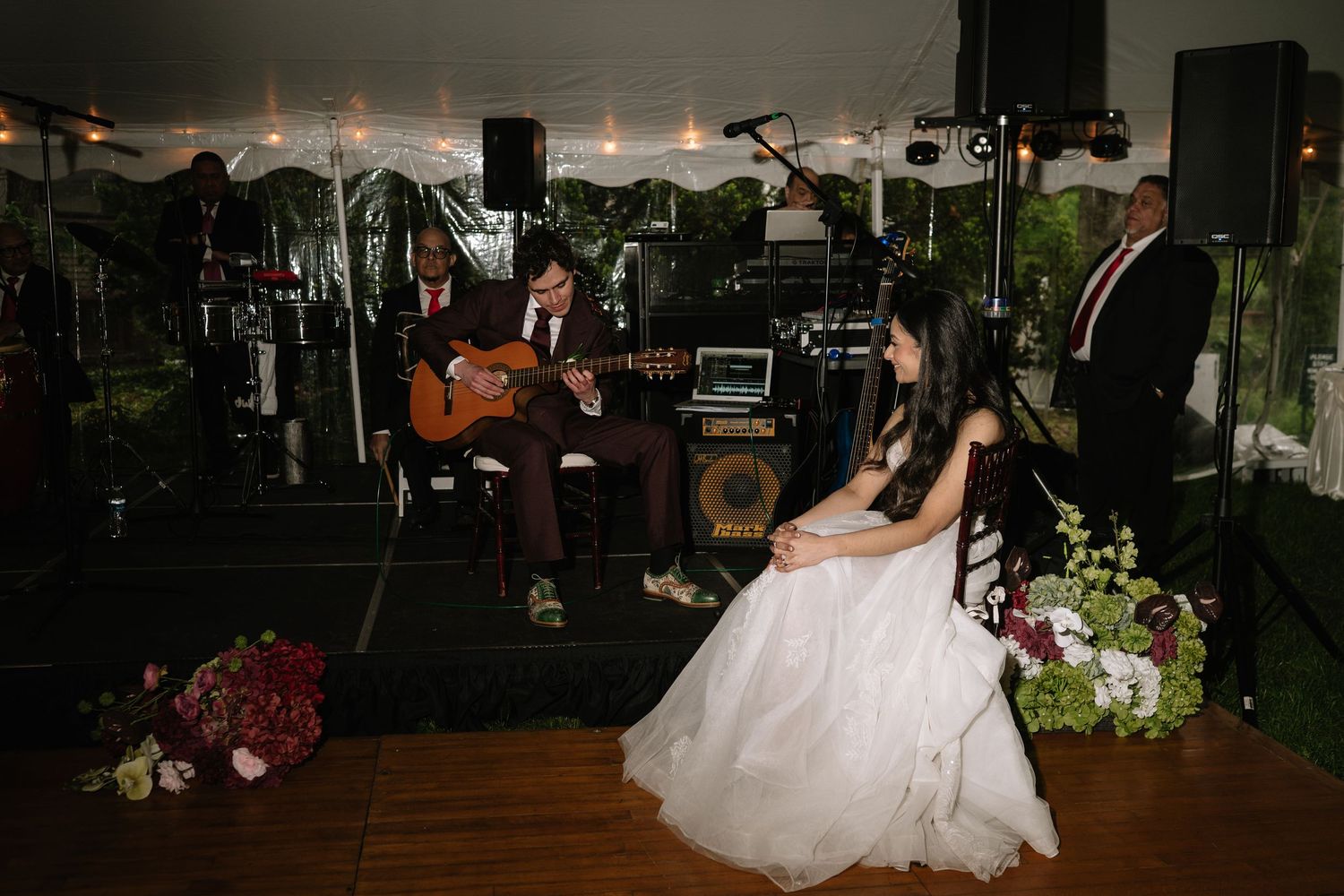 Bride in white gown seated at outdoor wedding reception while guitarist performs on stage under tent.