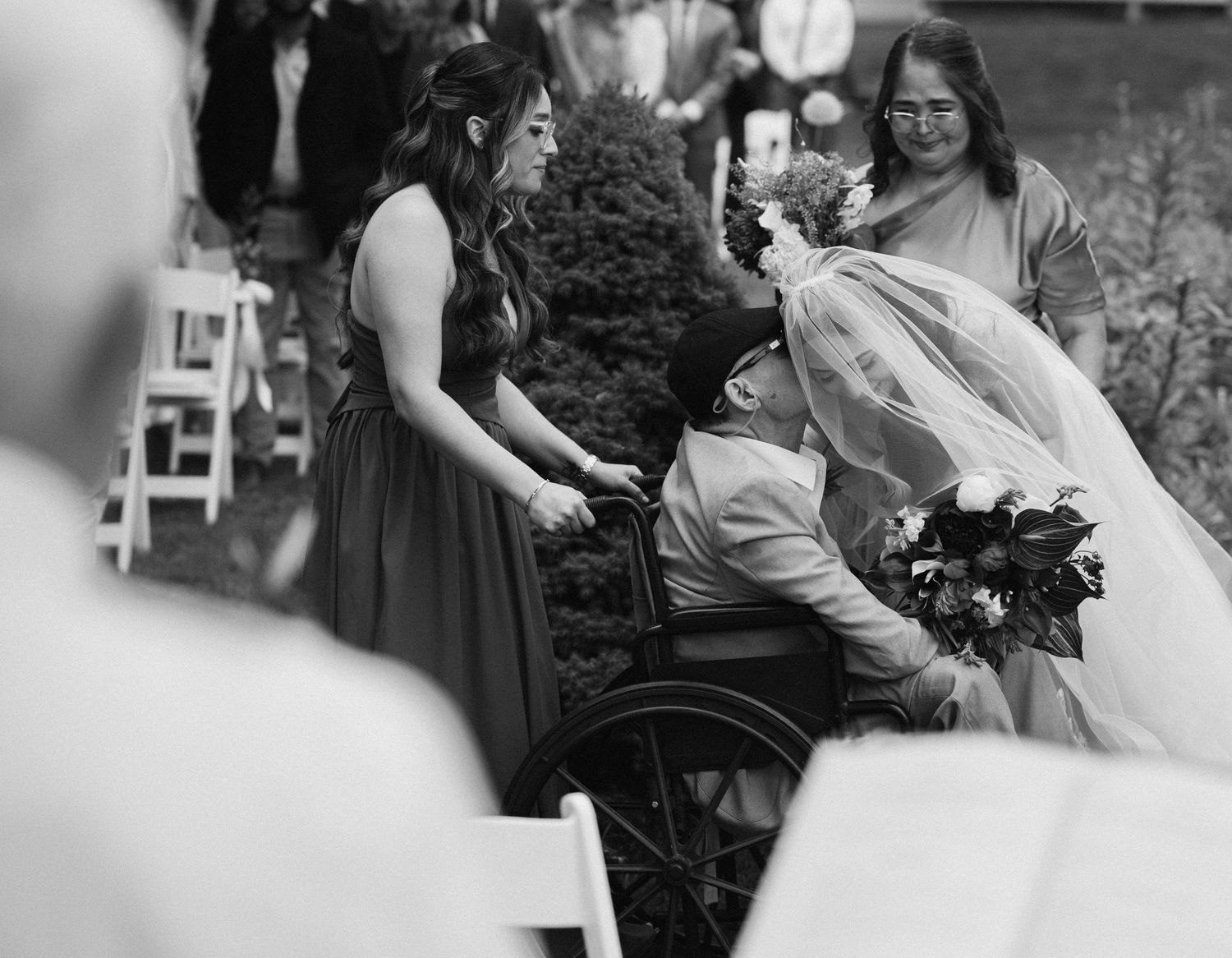 Black and white wedding photo of bride kissing man in wheelchair at outdoor ceremony, bridesmaid and guest nearby.