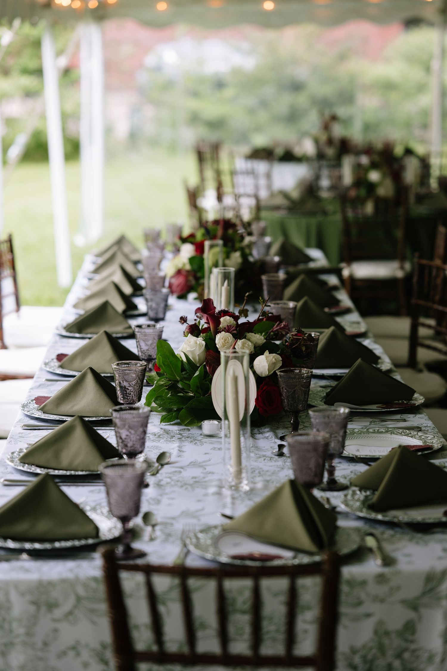 Elegant wedding reception table with green napkins, floral centerpieces, purple glasses, and white tablecloth under a tent.