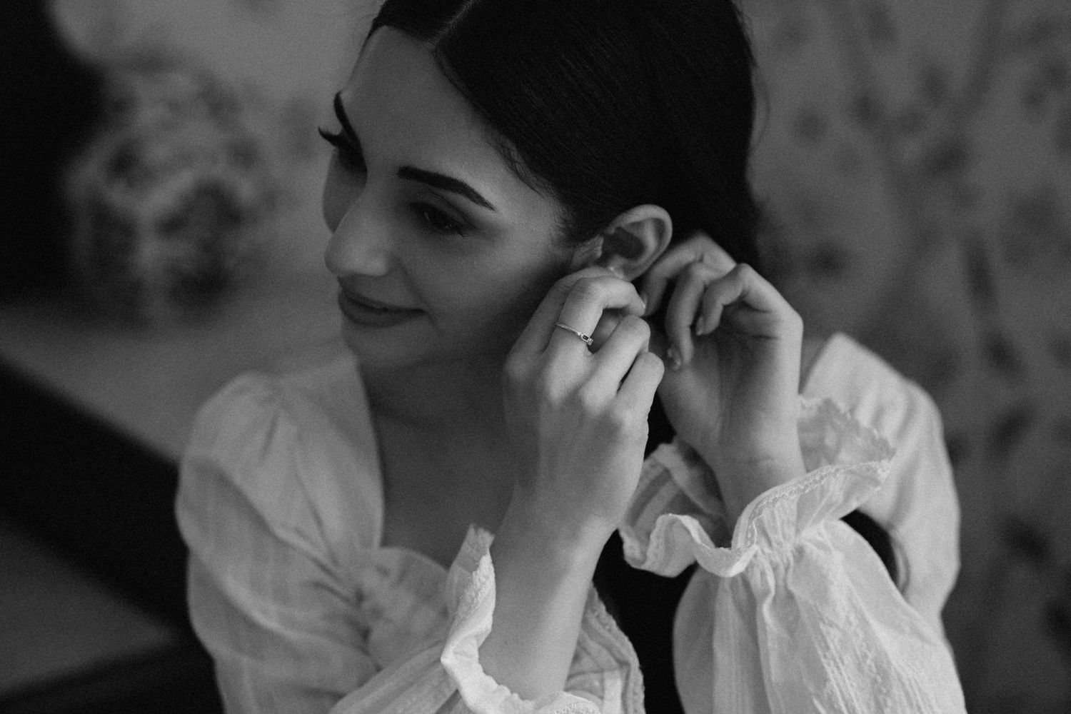 Black and white photo of a woman in a white ruffled blouse putting on earrings, wearing a ring.