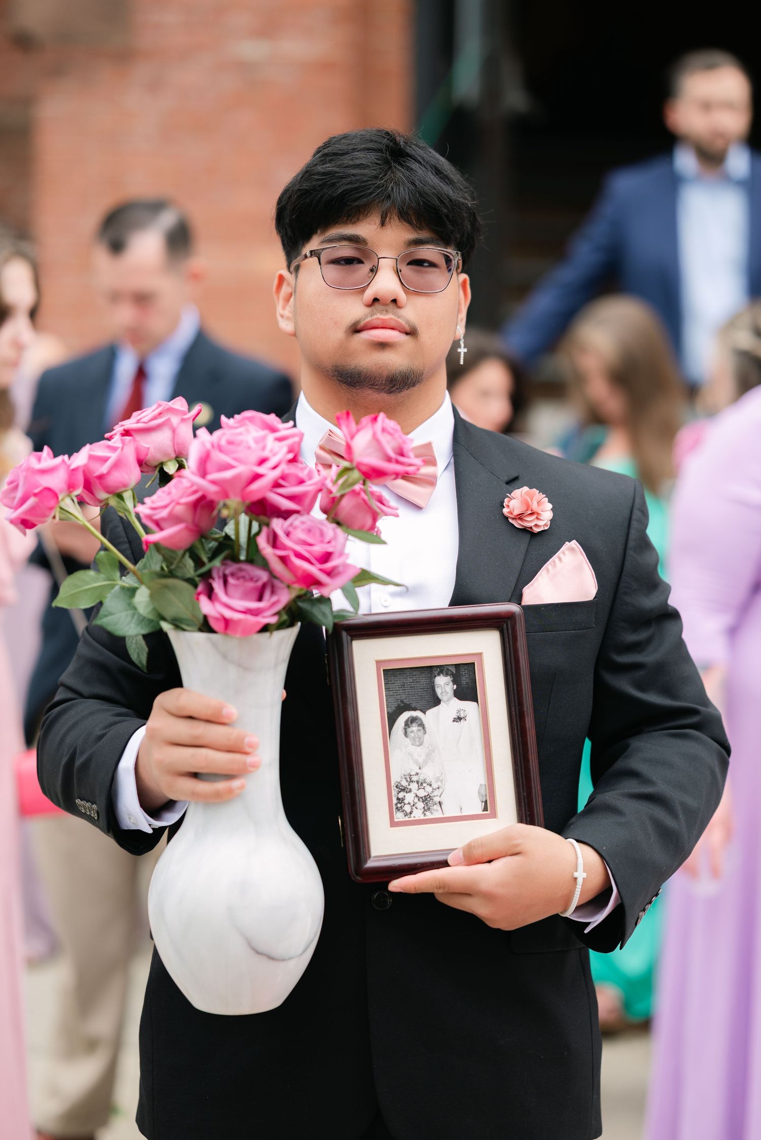 Groomsmen in black suits hold white vases with pink roses and framed photos during a wedding ceremony.