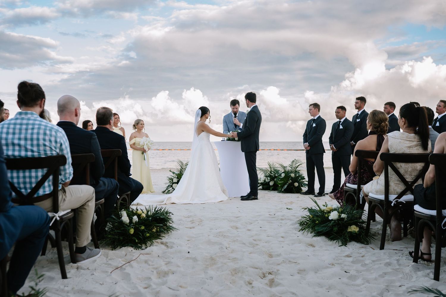 Beachfront wedding ceremony with guests seated on white chairs under cloudy skies.