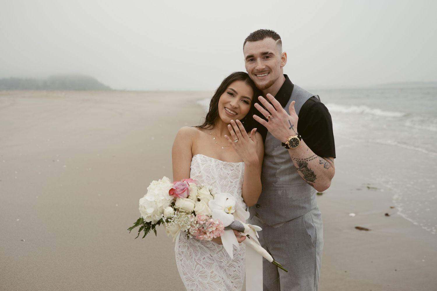 Wedding couple embracing on a misty beach with white floral bouquet and grey suit.