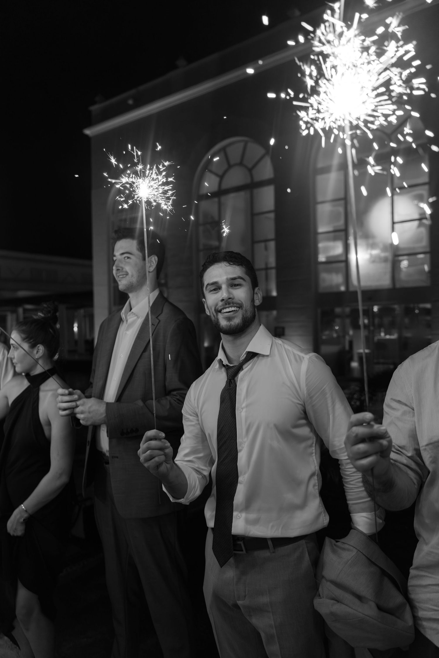 Wedding guests celebrate with sparklers at night, creating a festive atmosphere outside a venue.
