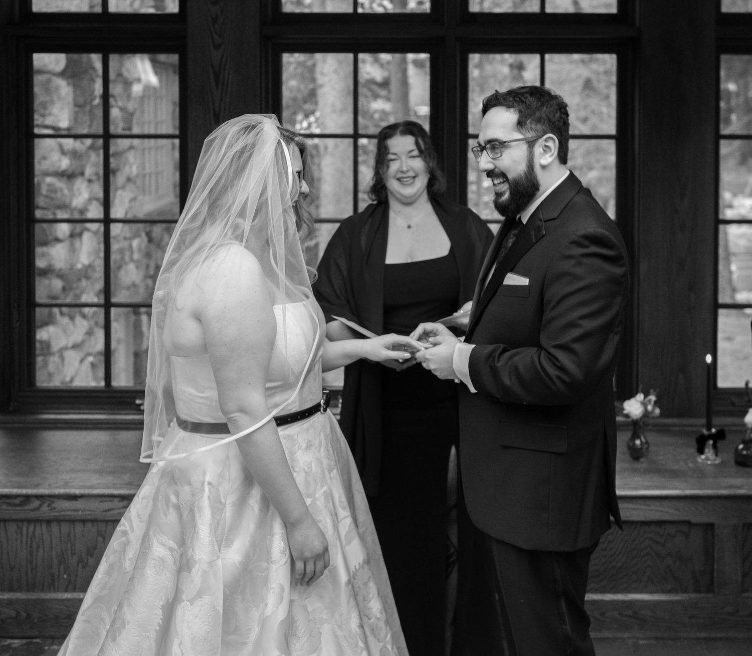 Bride and groom exchange rings at wedding ceremony as smiling officiant watches in black and white photo.