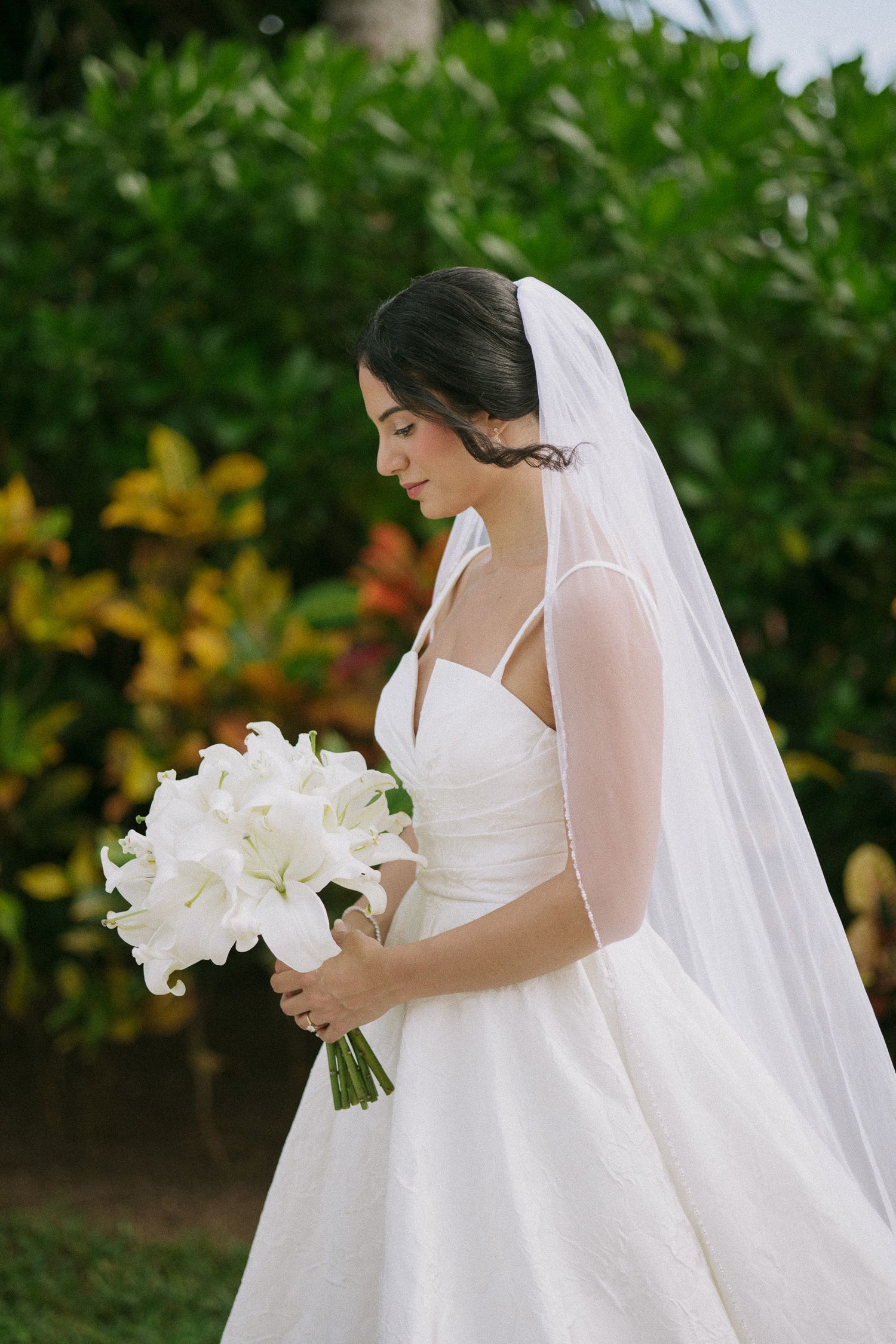 Elegant bride in white dress holding bouquet of white lilies against garden backdrop.
