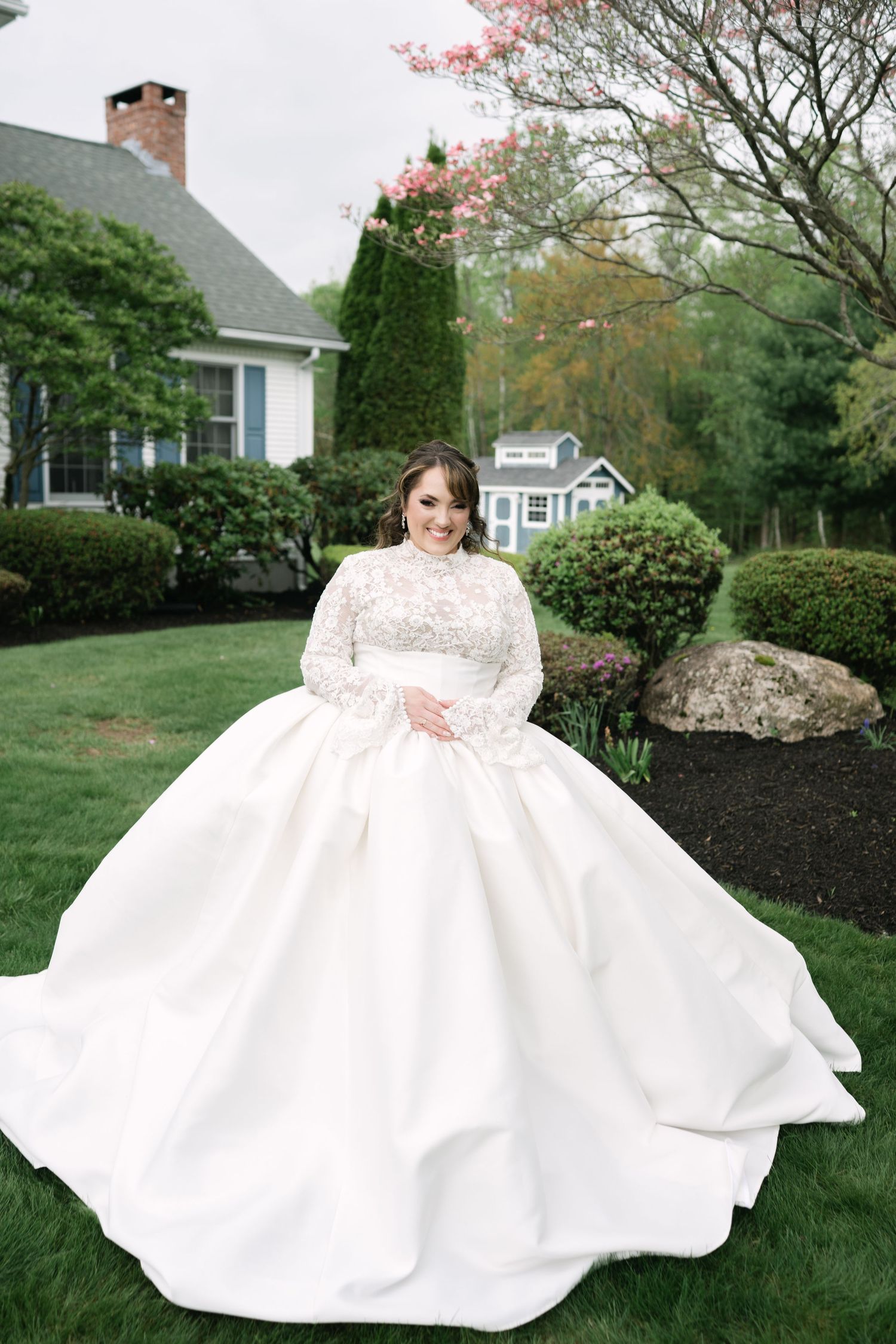 A bride in a flowing white gown poses gracefully on a lawn with blooming spring trees.