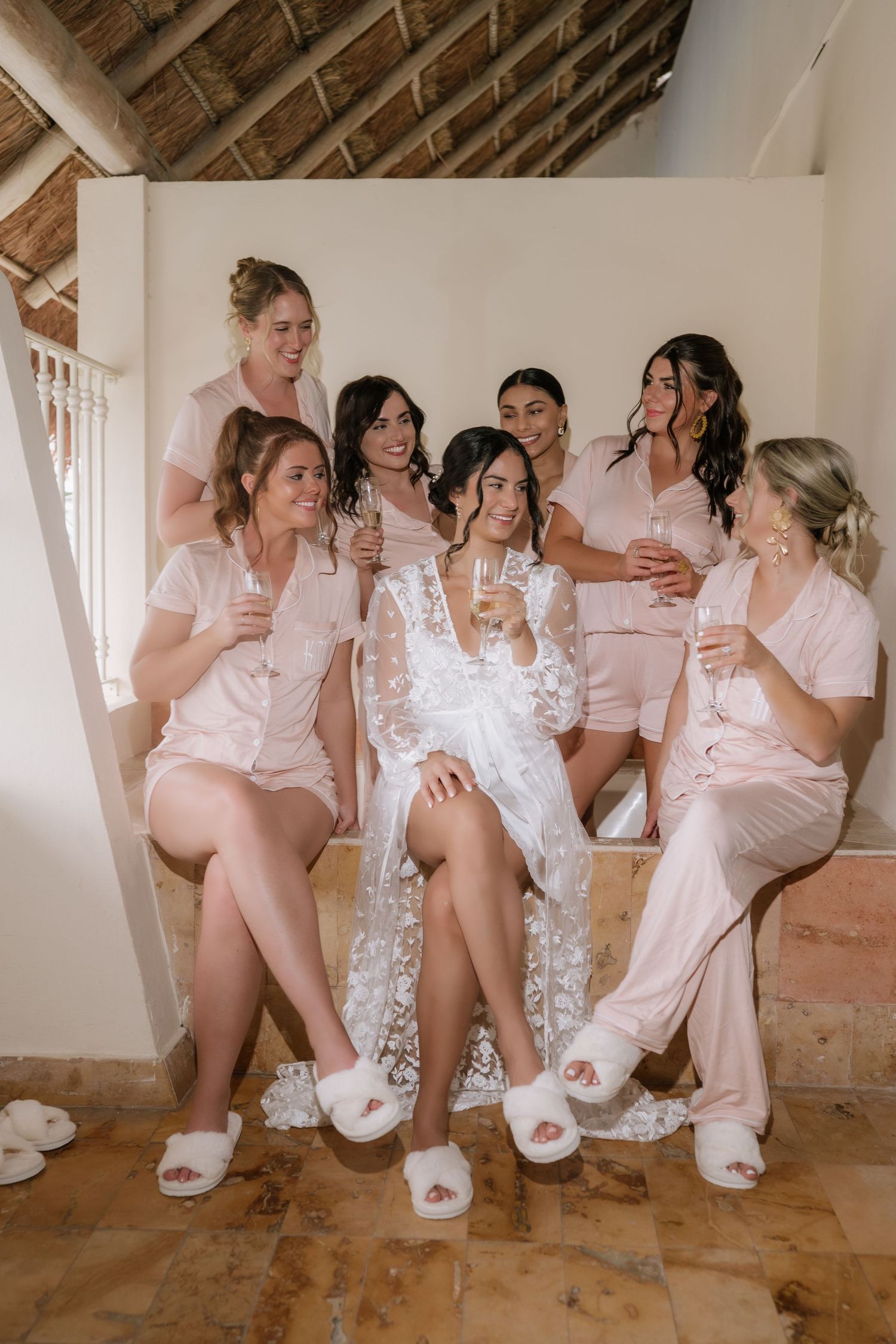 A bridal party in pink pajamas and white slippers share champagne together during wedding day preparations.