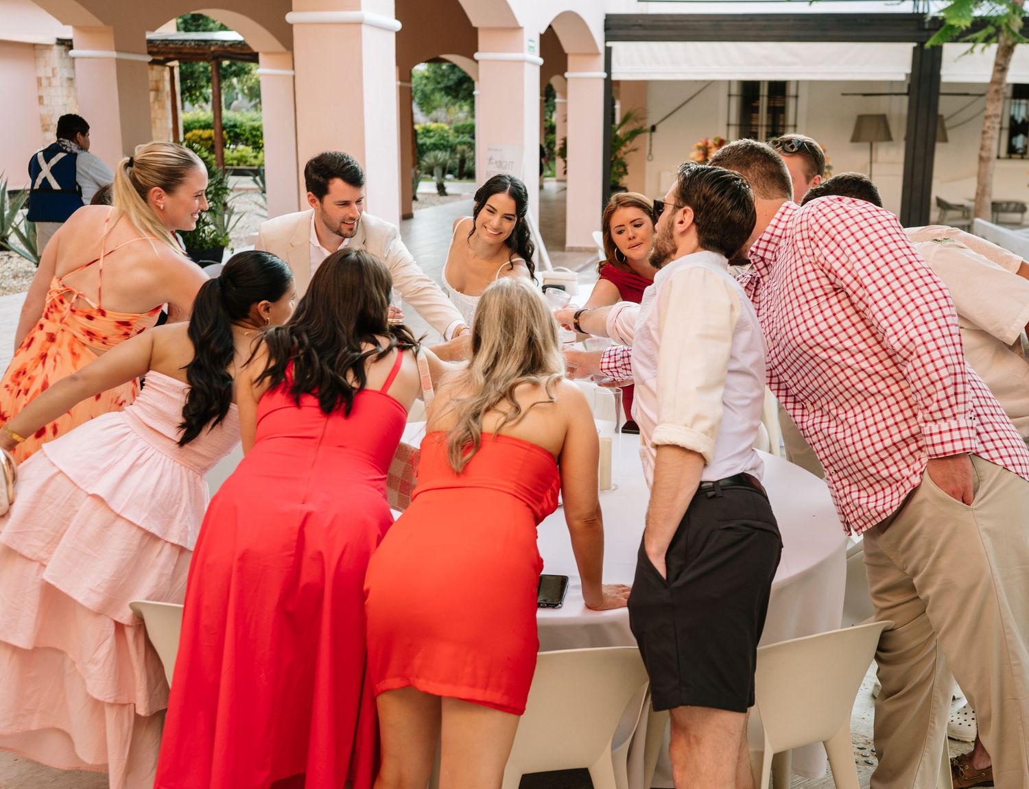 A group of wedding guests huddle together laughing and socializing at an outdoor venue with arched columns.