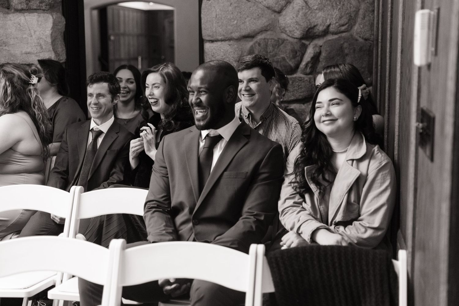 Black and white photo of smiling wedding guests seated in white chairs at an elegant indoor ceremony.