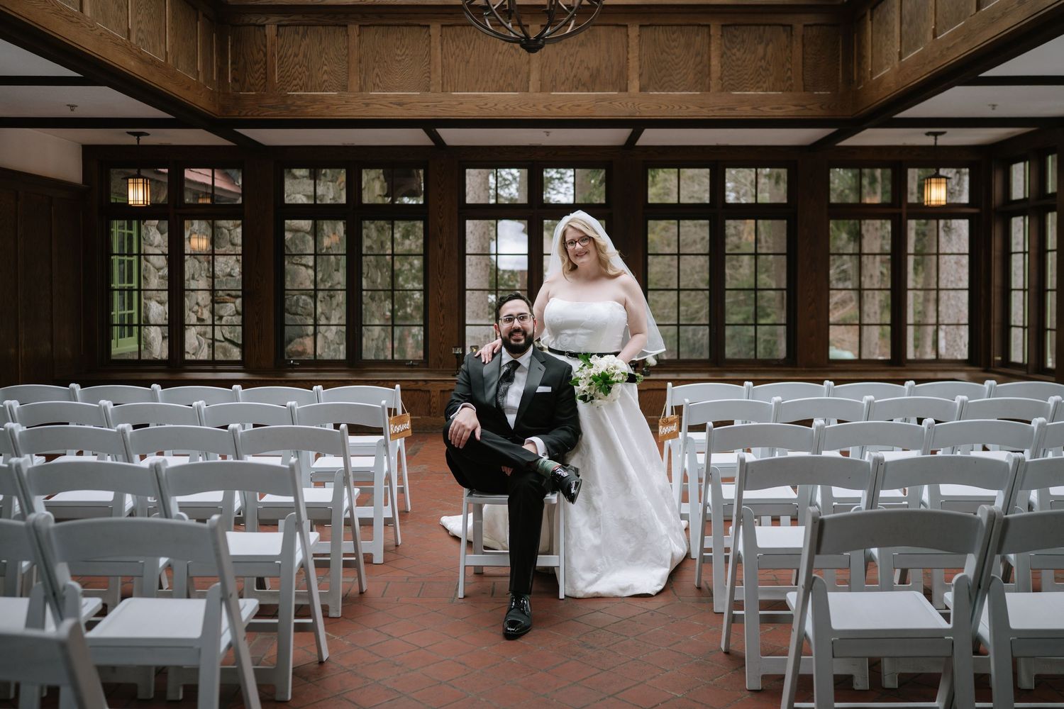 Bride and groom pose in elegant wood-paneled wedding venue with white folding chairs before ceremony.