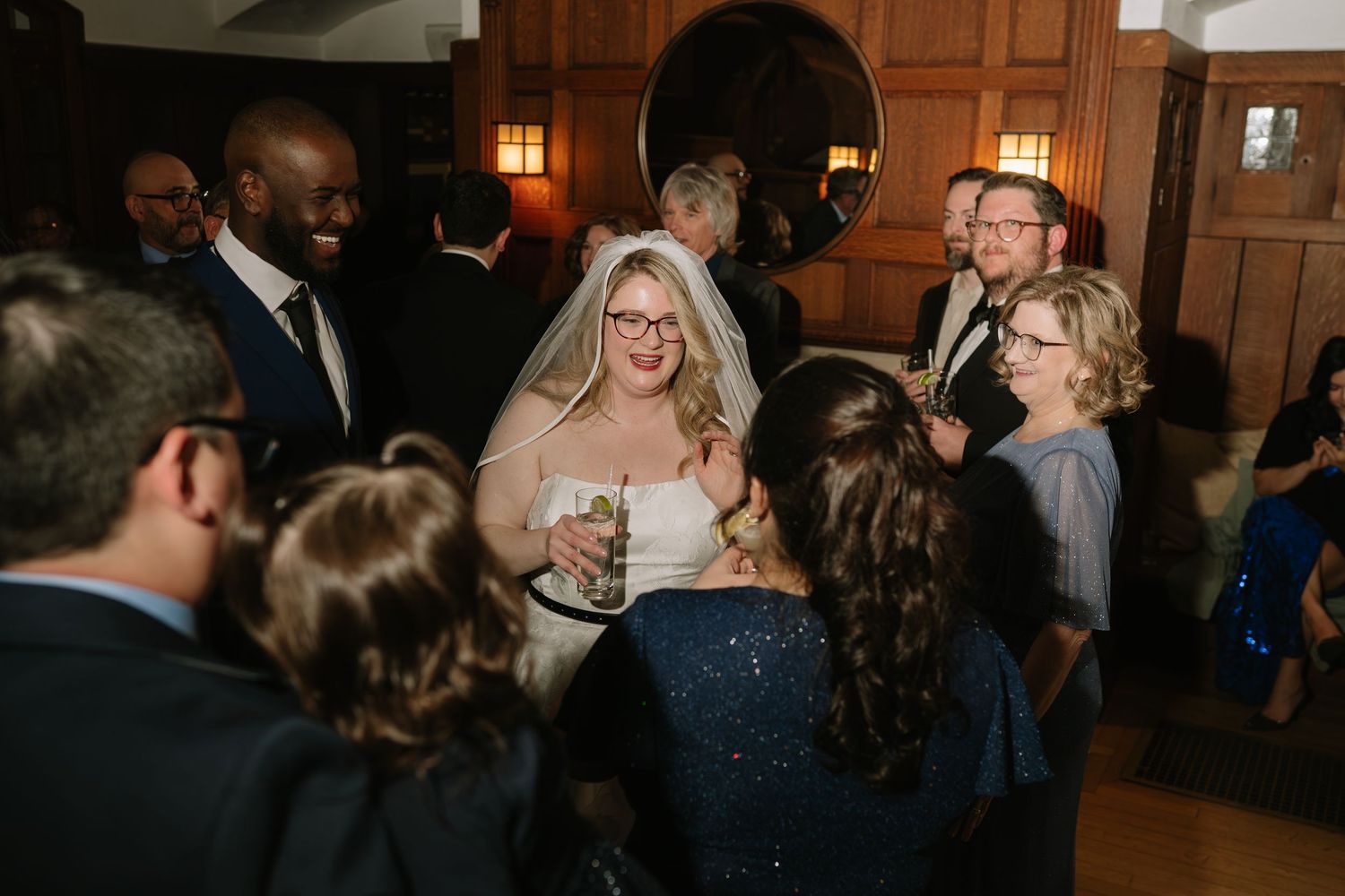 A bride in a white gown and veil mingles with guests at a wedding reception in a wood-paneled venue.