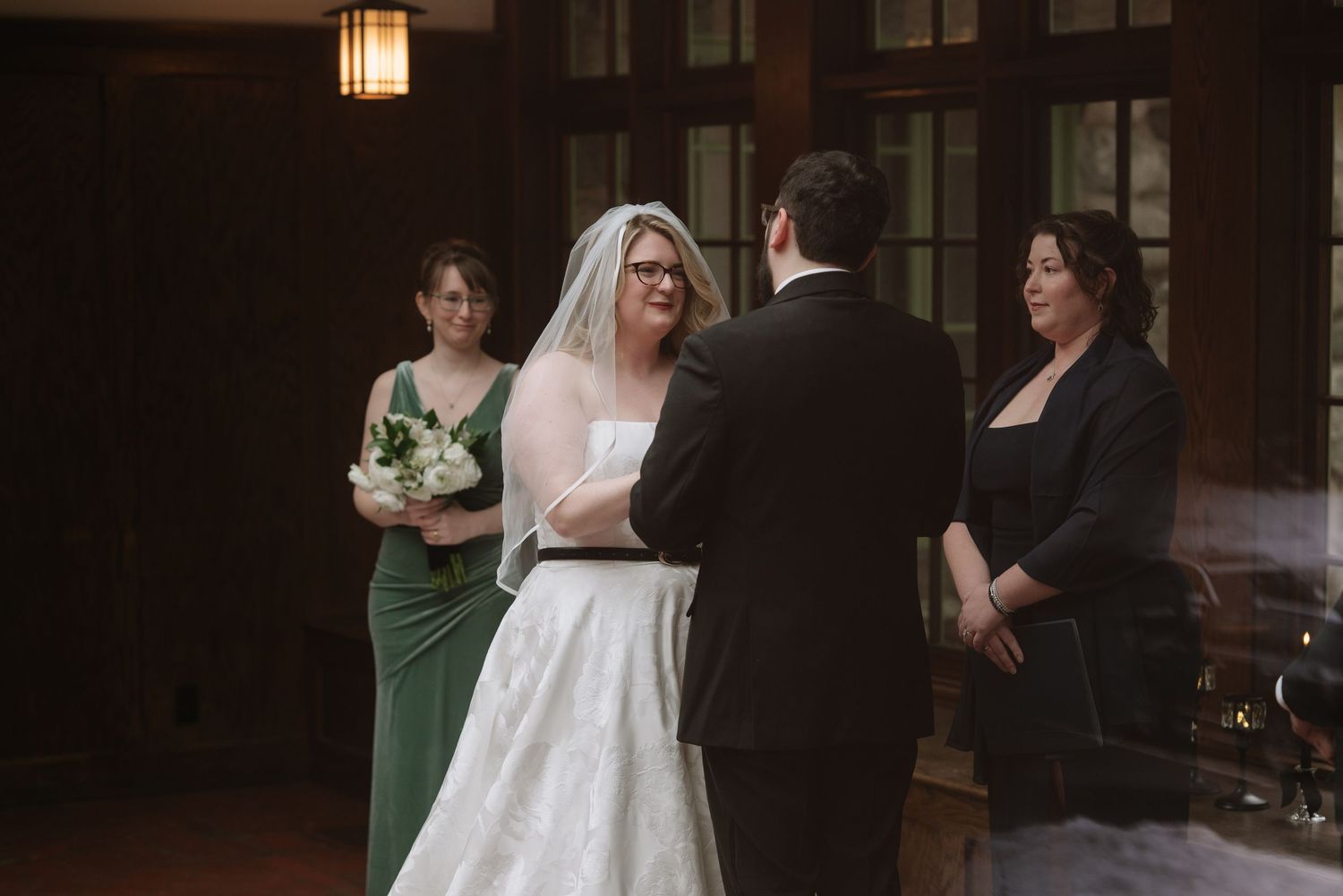 Bride and groom exchange vows at indoor wedding ceremony with bridesmaid in green dress and officiant nearby.