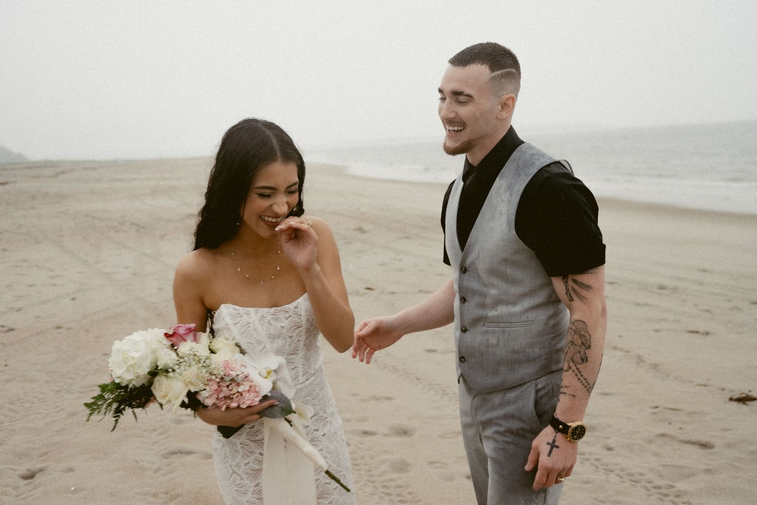 A couple in wedding attire shares an intimate moment on a cloudy beach.
