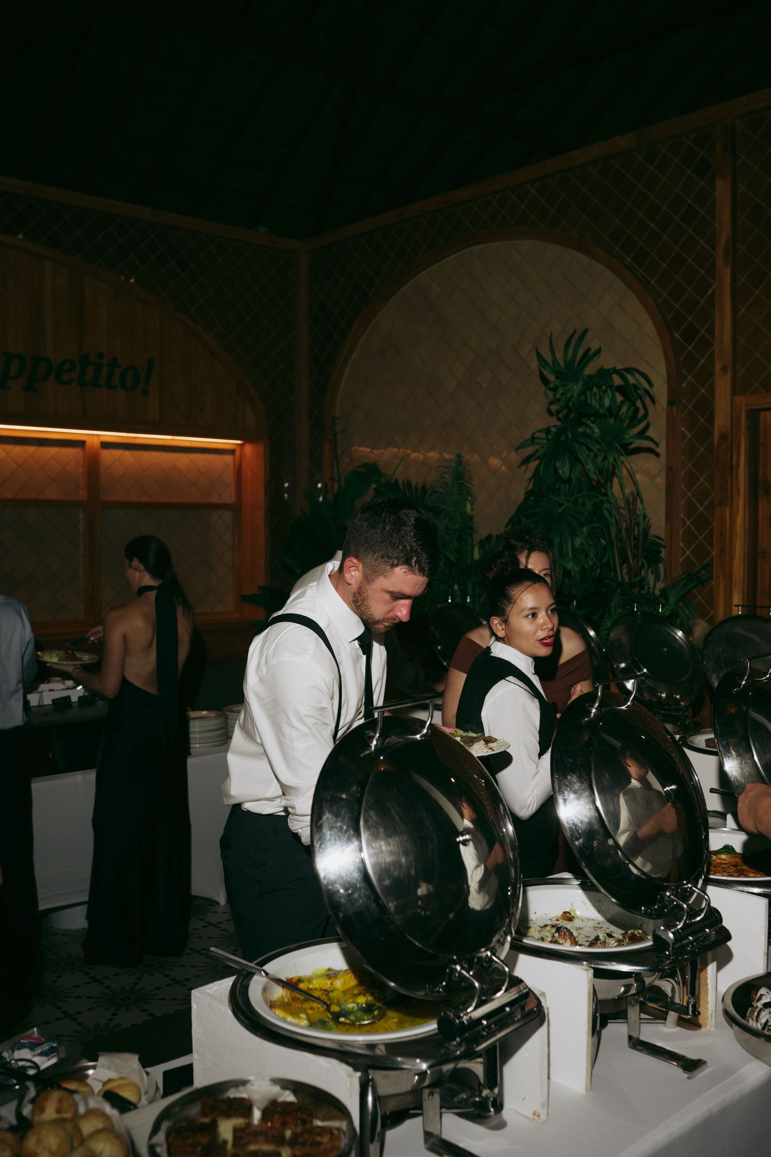 Catering staff in white uniforms serve food from covered silver serving trays at an event.