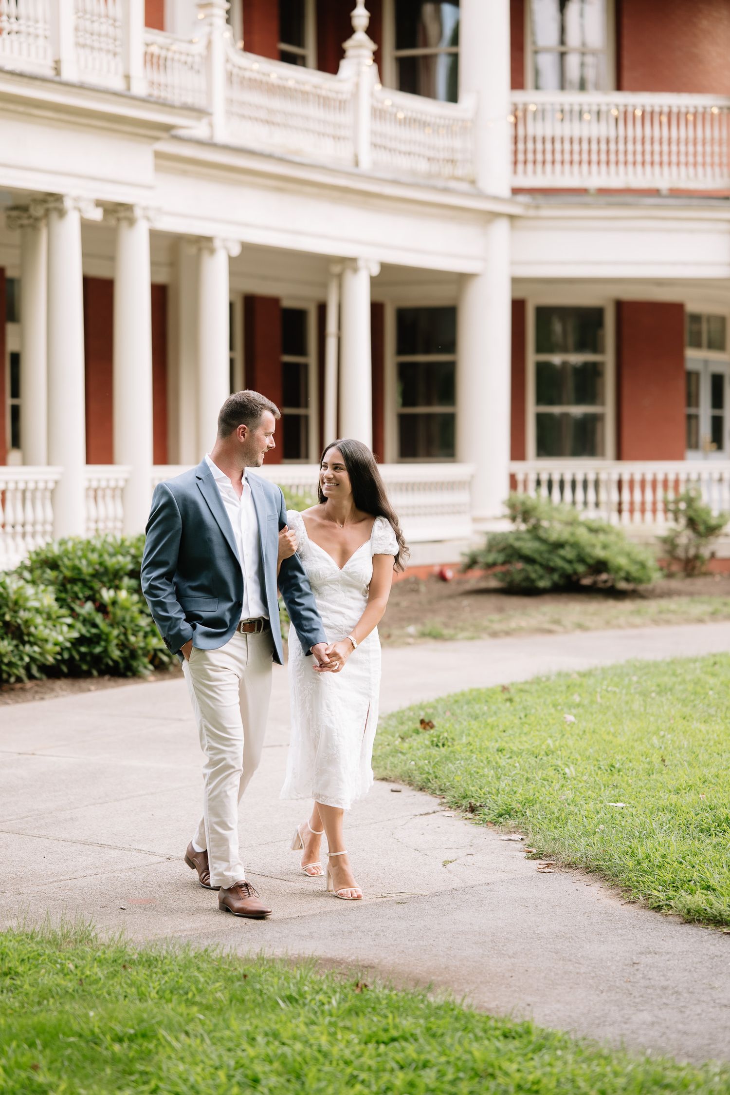 A couple in white and gray outfits walk playfully along a historic building's colonnade.