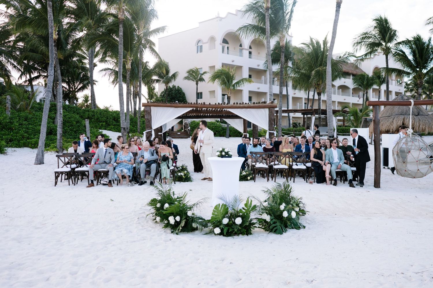 A beachfront wedding ceremony setup with white chairs and palm trees along a tropical shoreline.