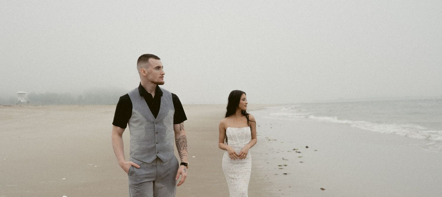 A couple walks along a misty beach, with the person in a gray vest and the other in a white dress.
