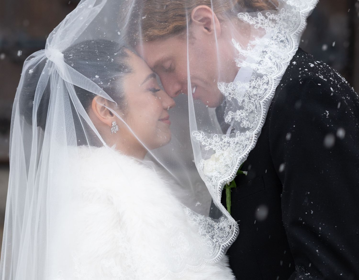 A romantic winter wedding moment captured under a delicate veil with snow falling.