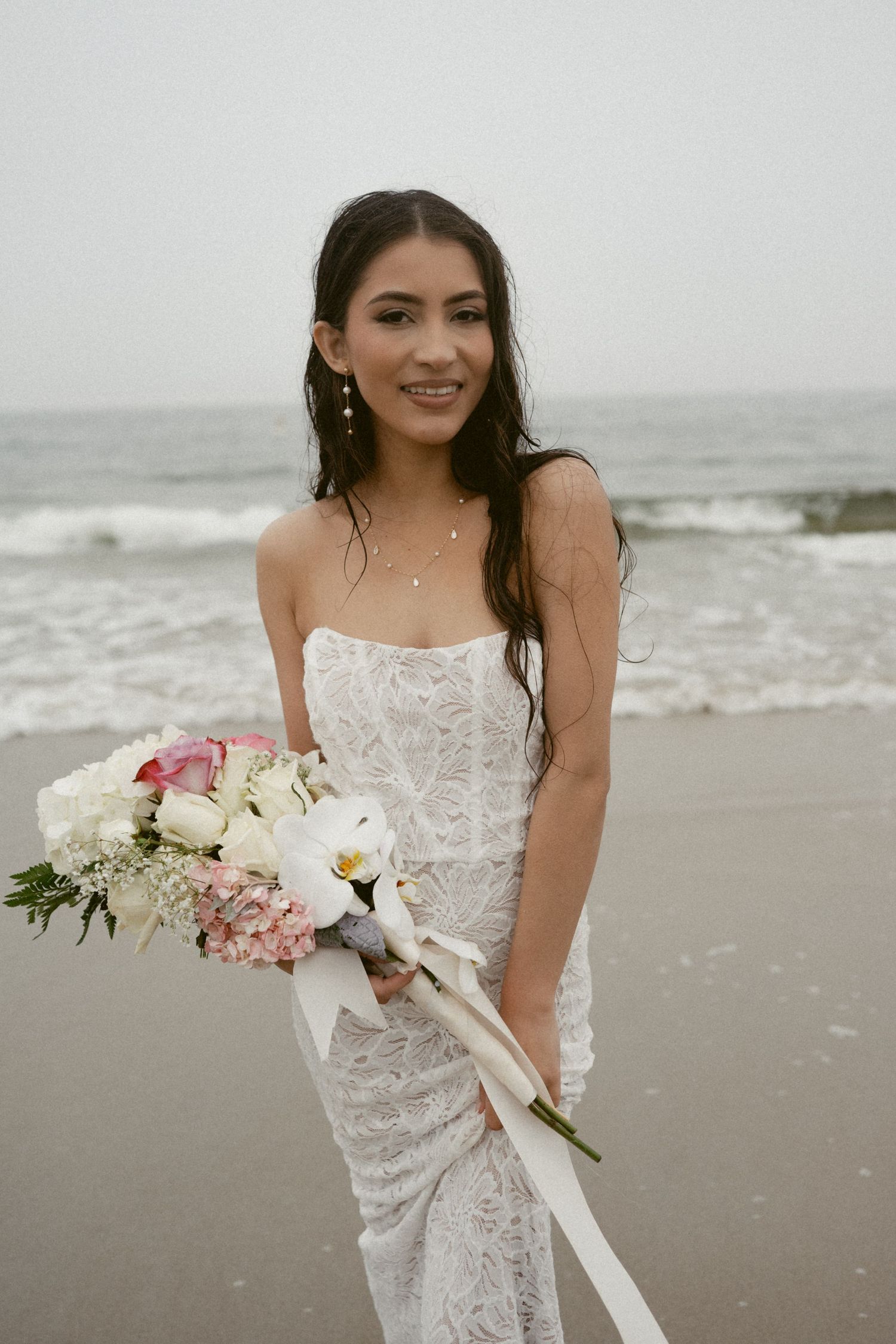 A delicate lace wedding dress is showcased against a moody beach backdrop with waves and grey skies.