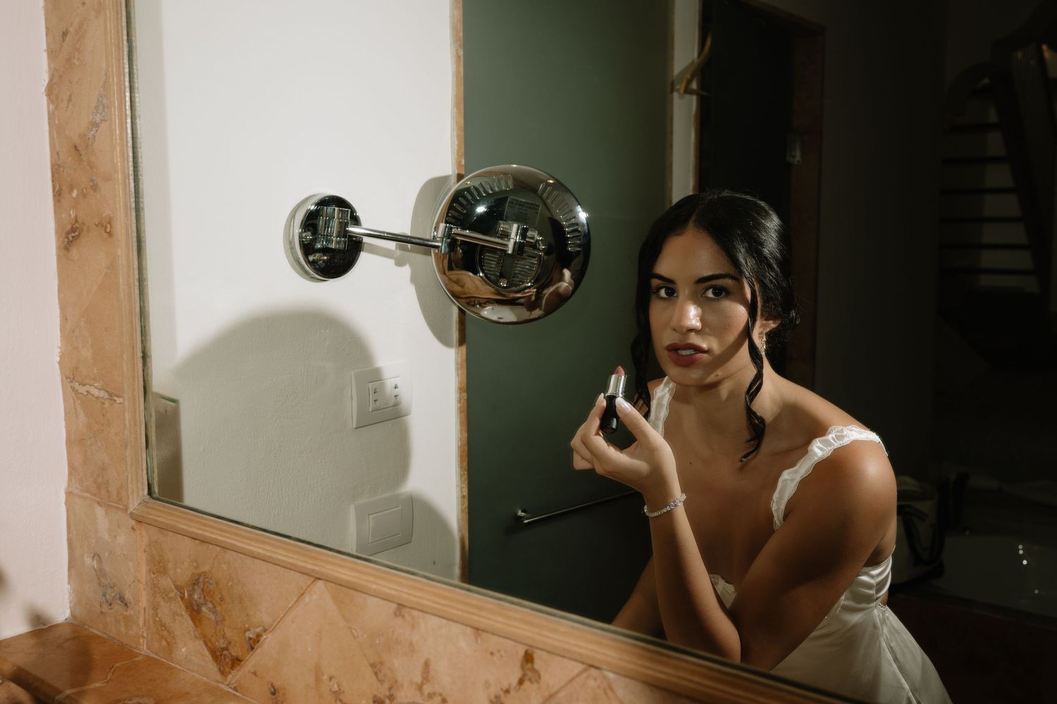 A person applying makeup in front of an illuminated bathroom mirror with a wall sconce.
