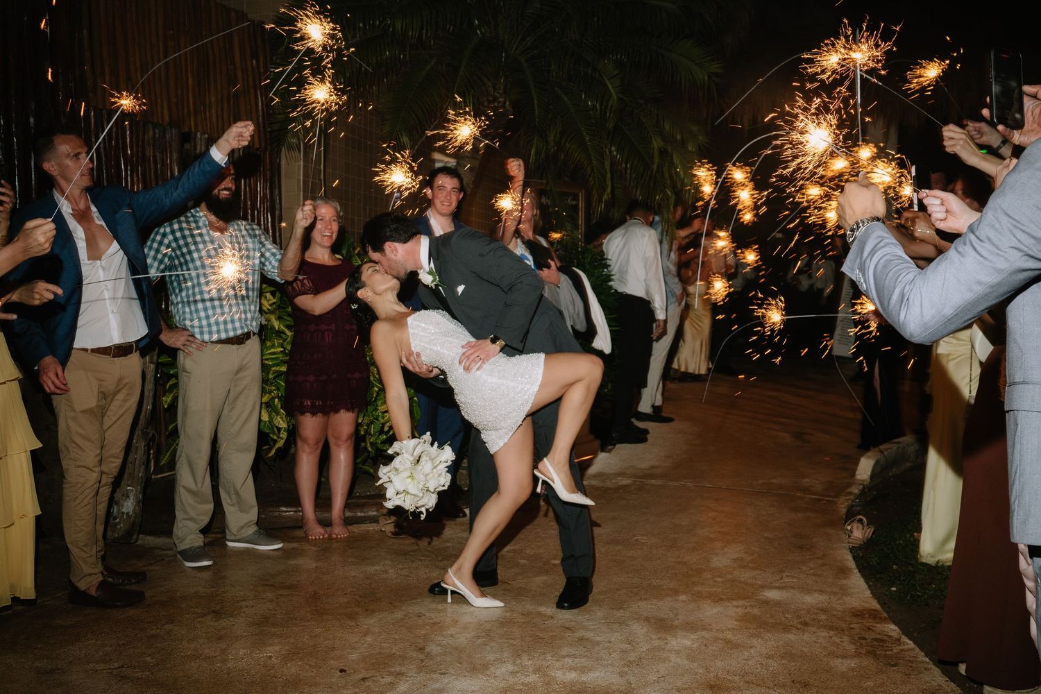 Wedding guests holding sparklers while celebrating as couple performs a dip dance move on outdoor patio.