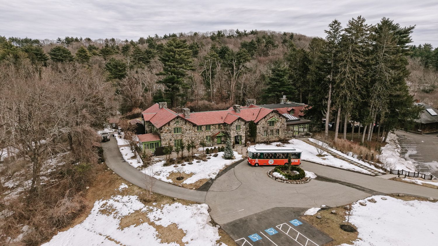 Aerial view of a stone lodge with red roof surrounded by snow-dusted grounds and pine forests in winter.