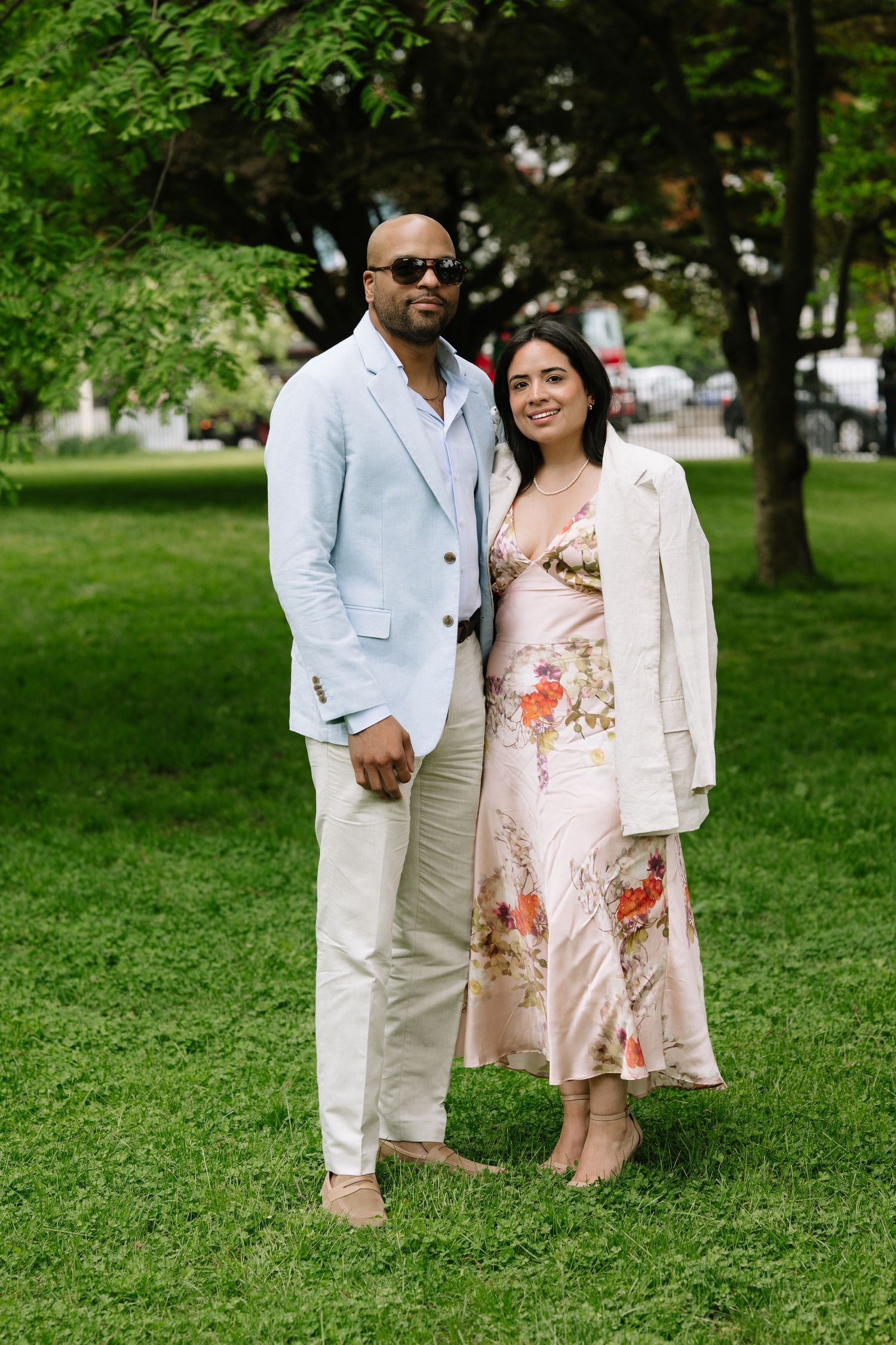 Stylish couple posing outdoors on green grass, man in light blue blazer, woman in floral midi dress.