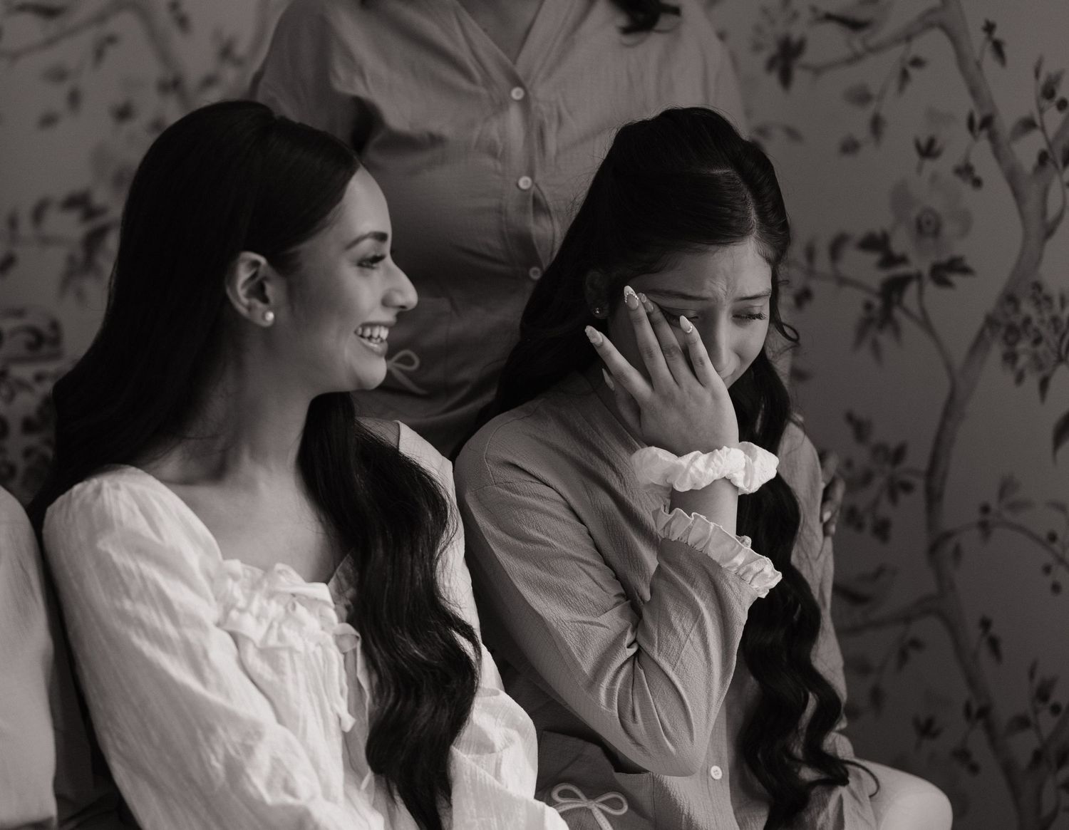 Two young women in white dresses sit together, one smiling and one covering her face, in a black and white photo.