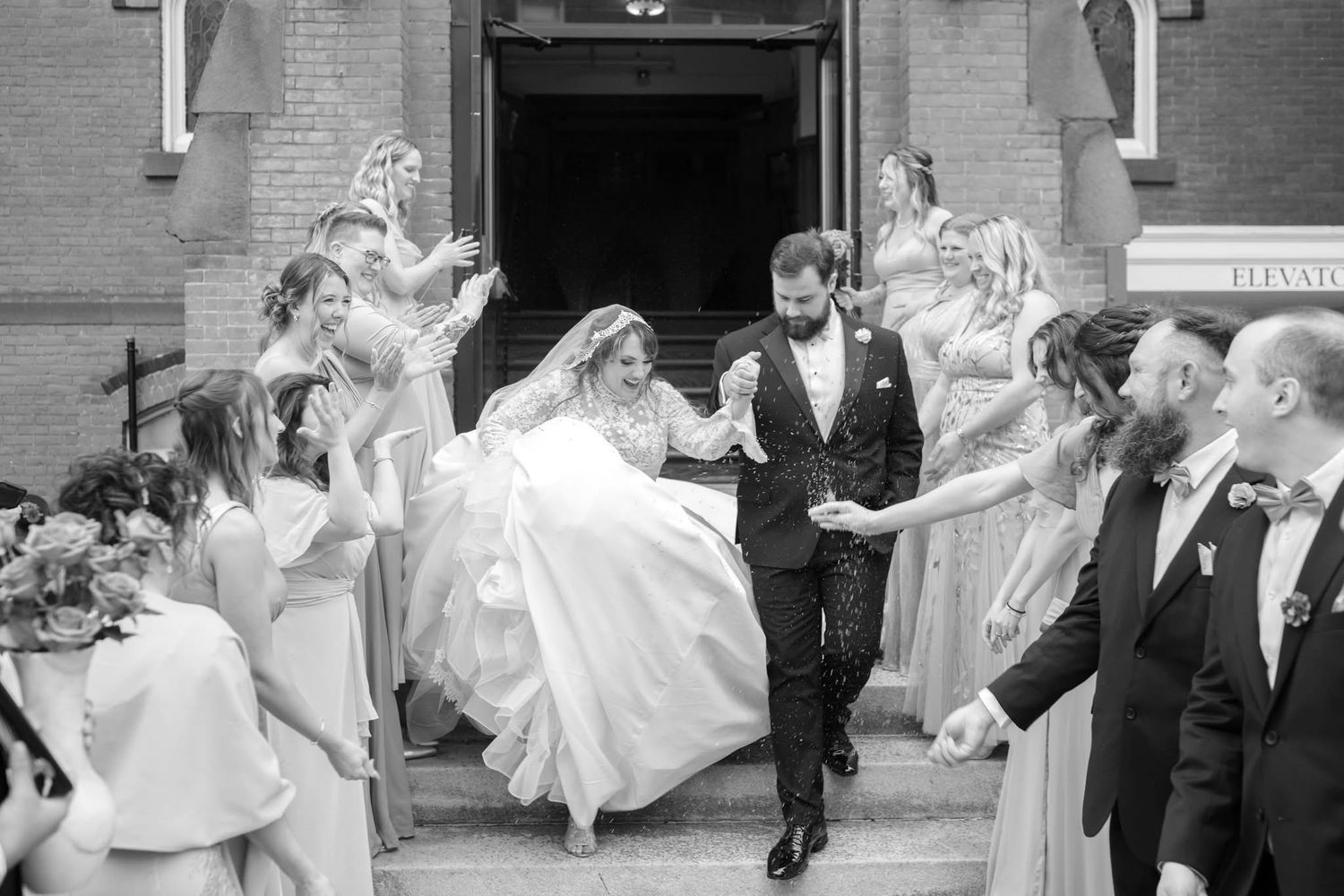 Wedding party dancing and celebrating in front of a brick building with joyful energy.