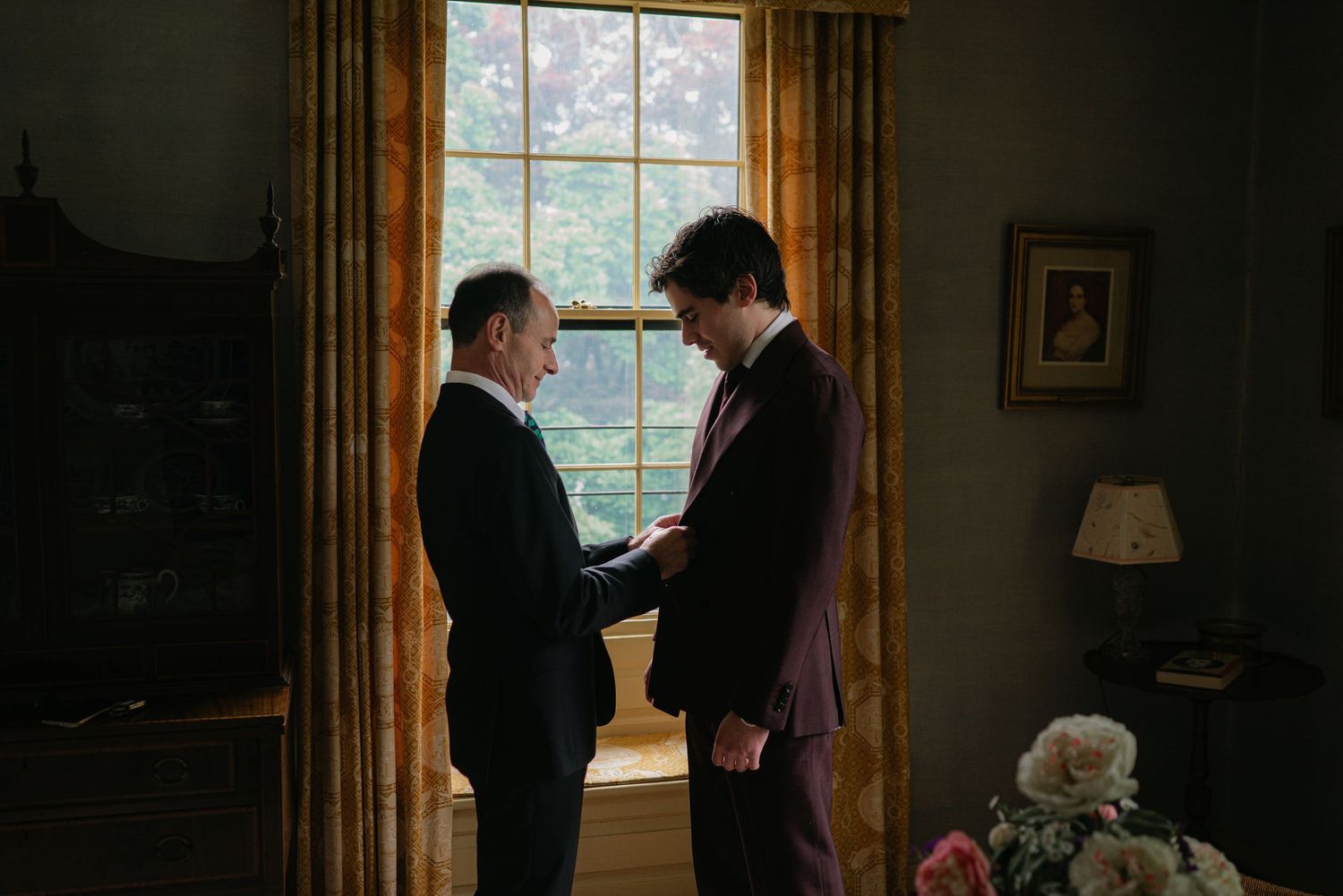 Two men in suits share a tender moment by a window, one adjusting the other's boutonniere before a wedding.