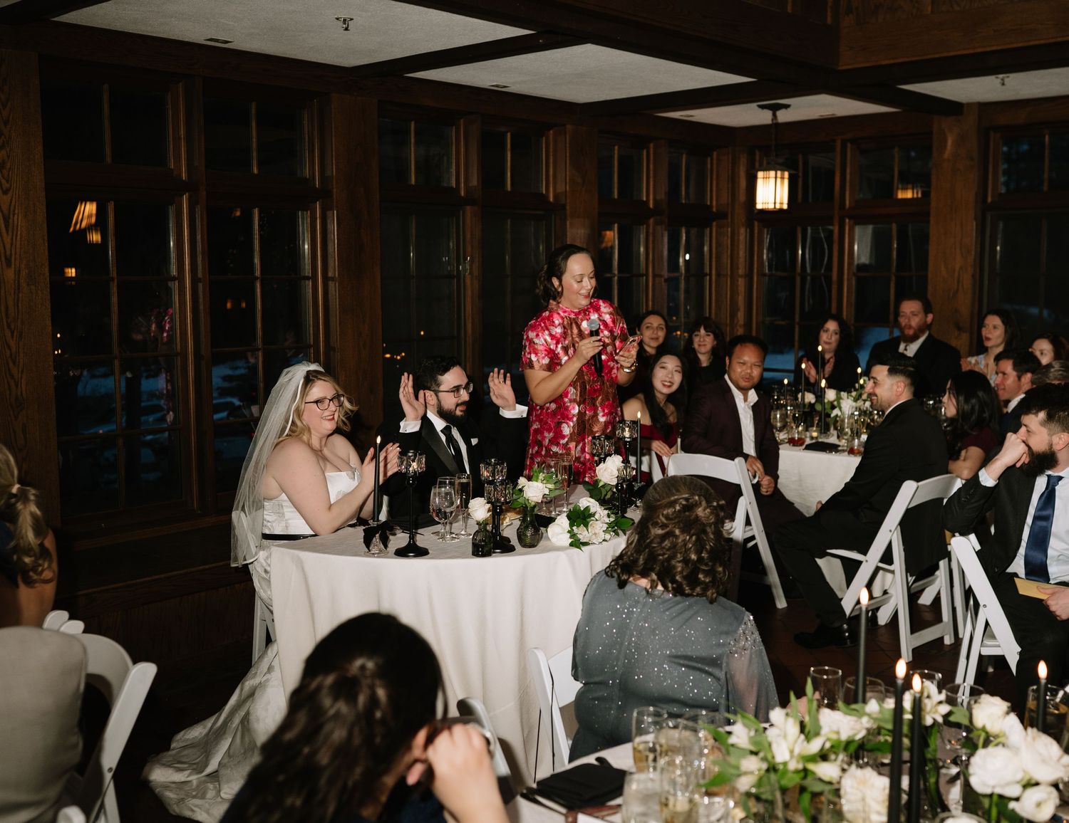 Woman in red dress giving a toast speech at an elegant wedding reception dinner with bride and guests seated at tables.