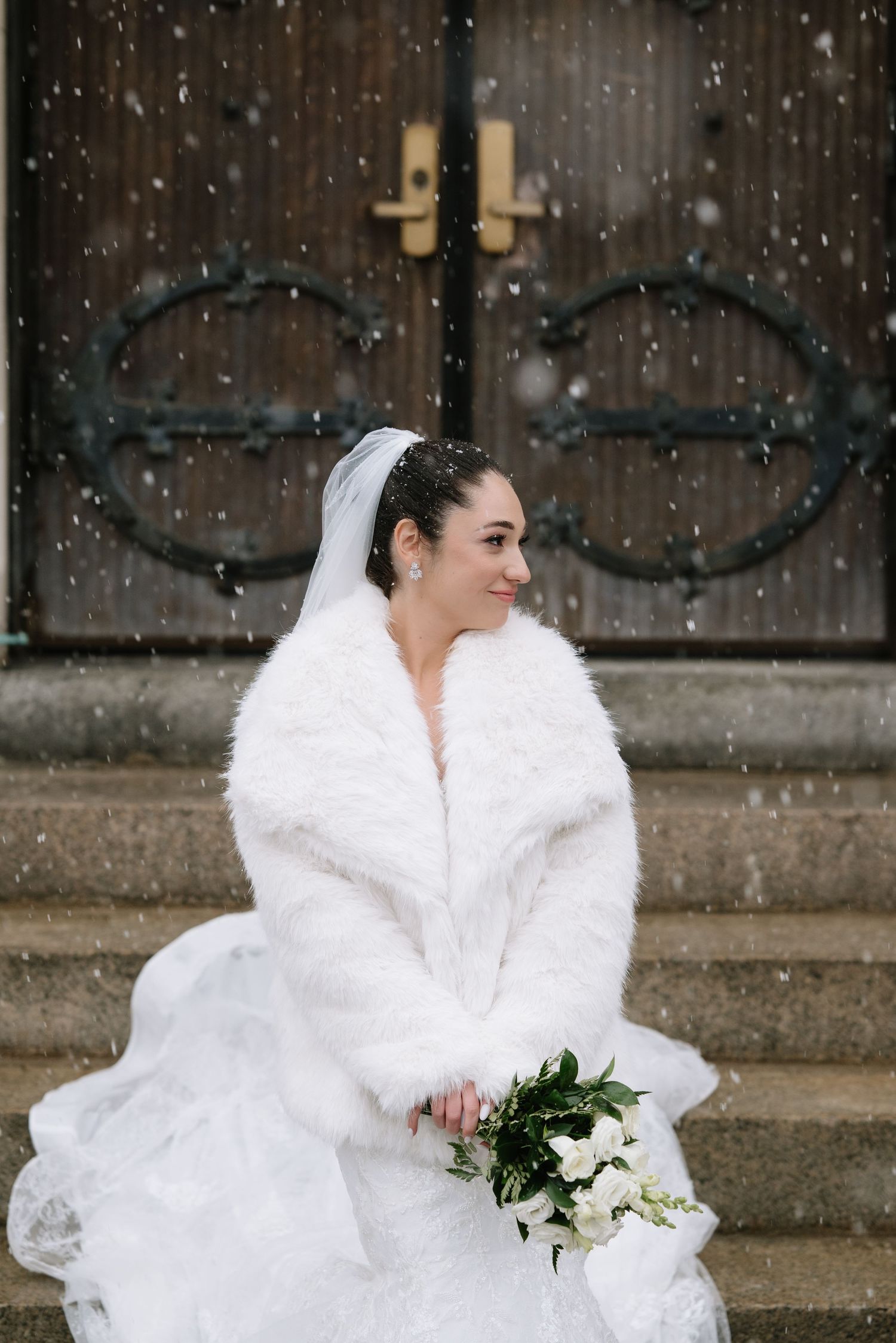 Bride in white fur wrap holds bouquet while snow falls outside church.