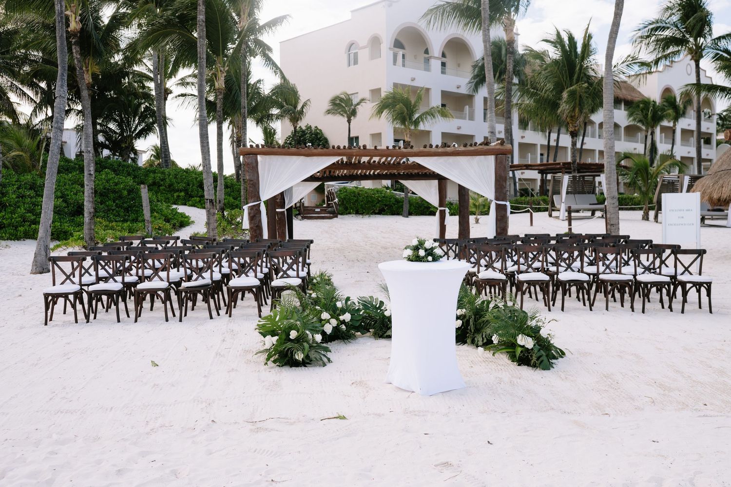 Elegant beach wedding setup with dark wooden chairs and white floral arrangements on a sandy shore by a white resort building.